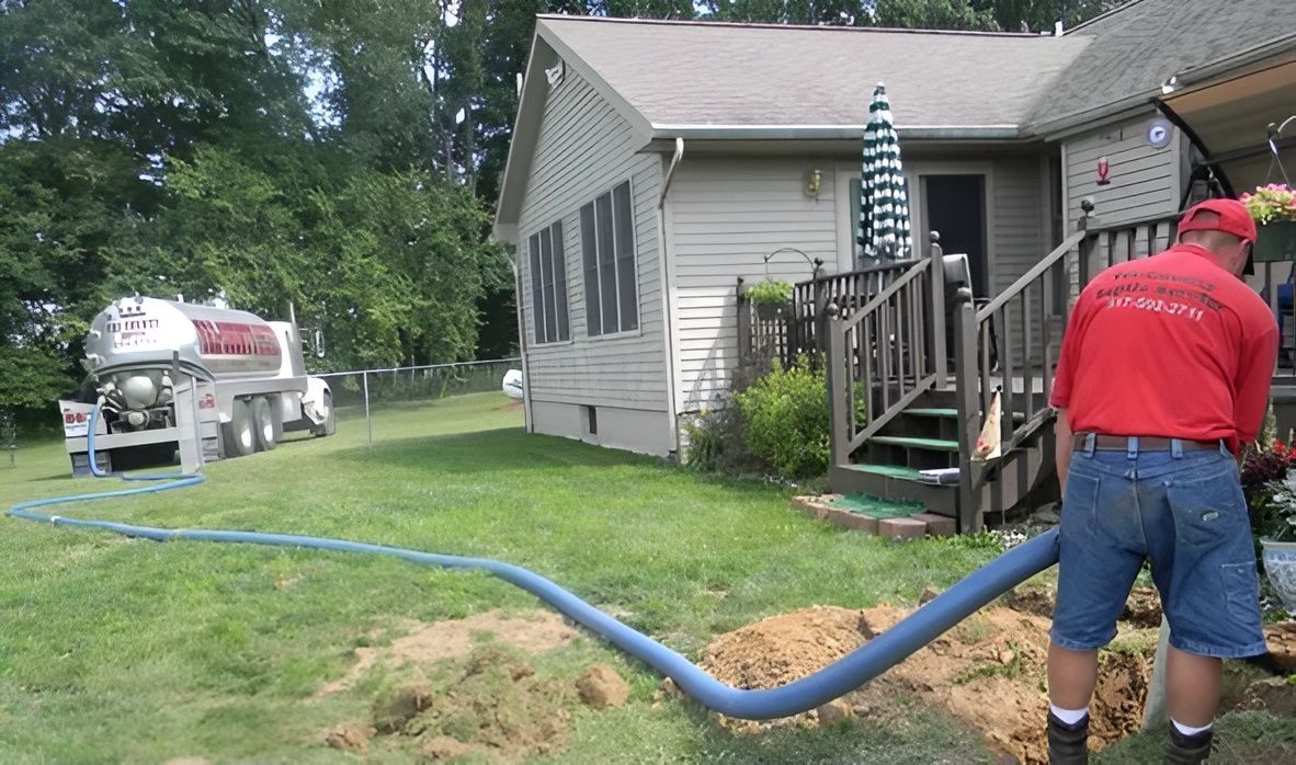 Man in red shirt working on a hose connected to a truck near a house.