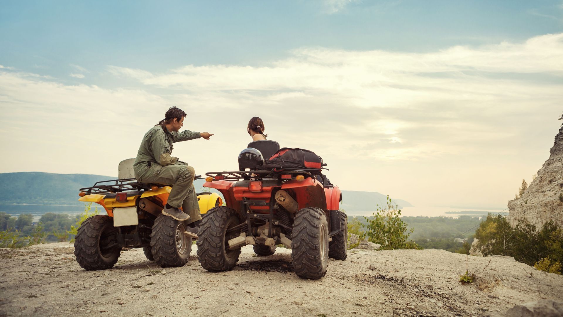 Two people on ATVs atop a hill, one pointing towards a distant landscape.