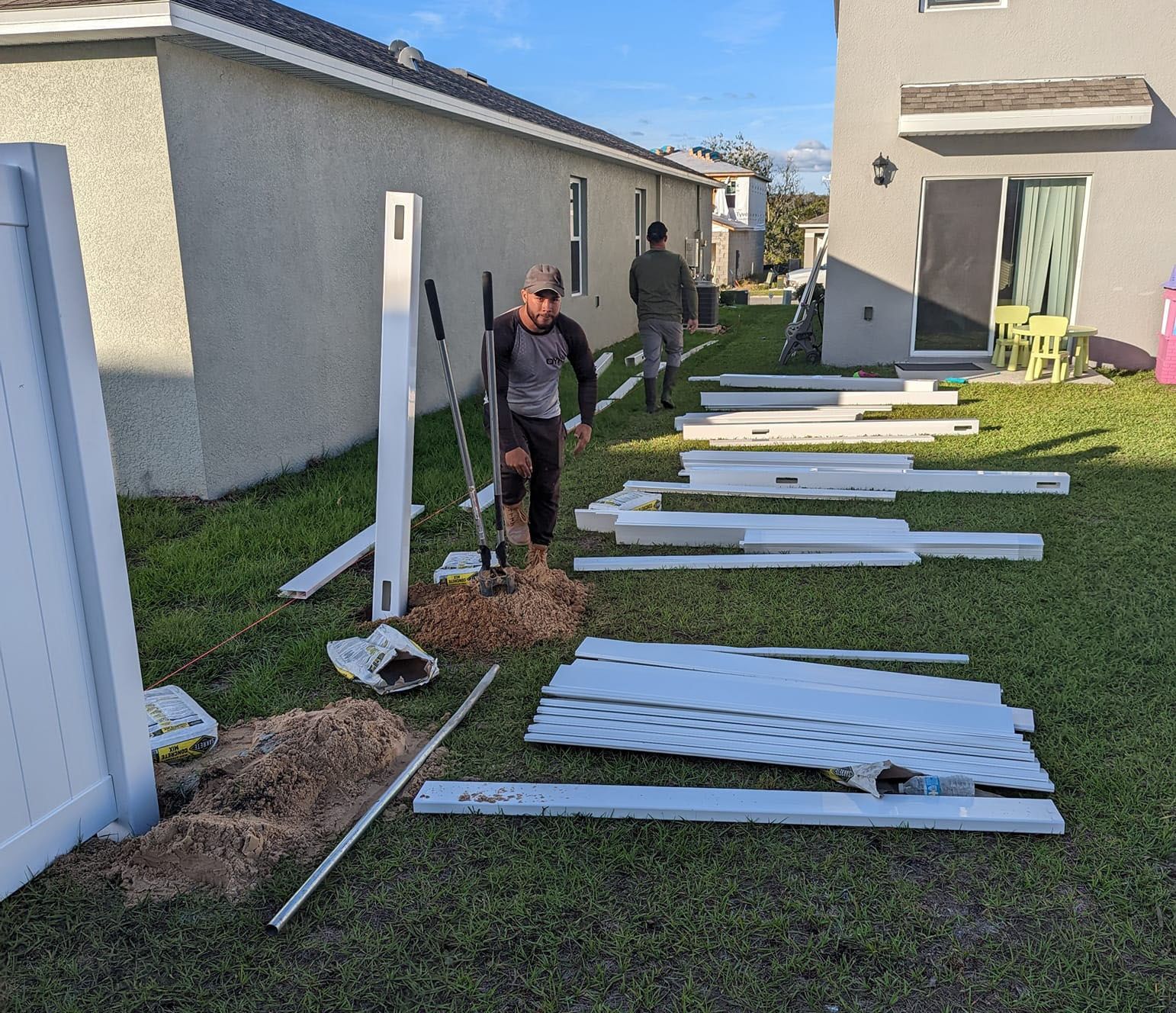 Two men installing a white vinyl fence in a grassy backyard. One digs, others assemble materials.