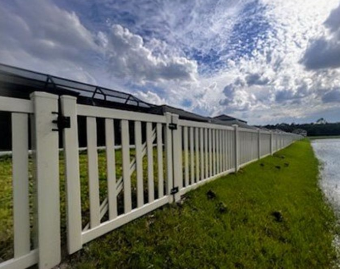 White picket fence along a green grassy area, near a lake, under a cloudy sky.