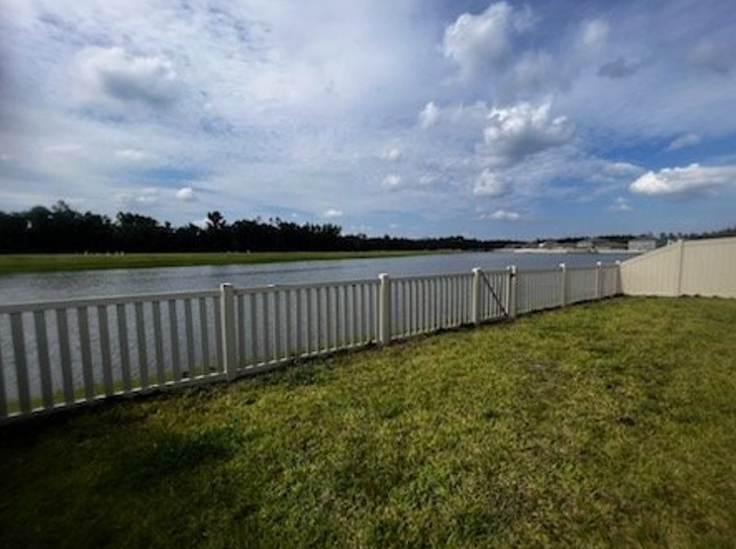 White fence along a body of water, green grass in foreground, cloudy sky.