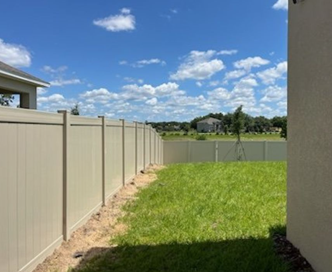 Tan vinyl fence surrounds a grassy backyard on a sunny day with blue sky and clouds.