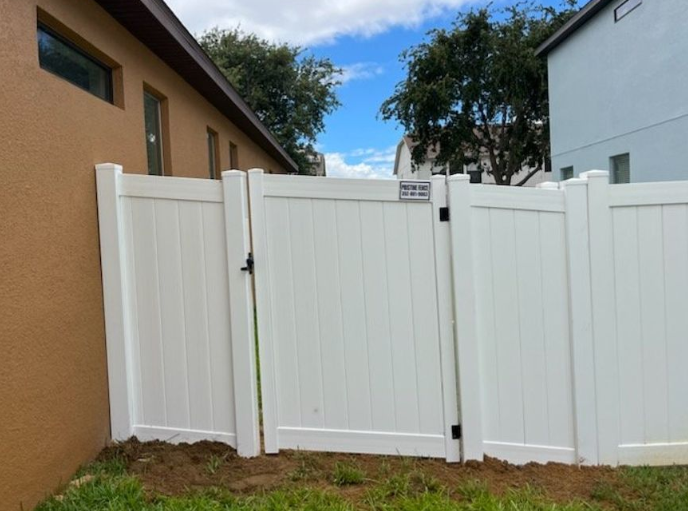 White vinyl fence with a gate between two buildings under a cloudy blue sky.