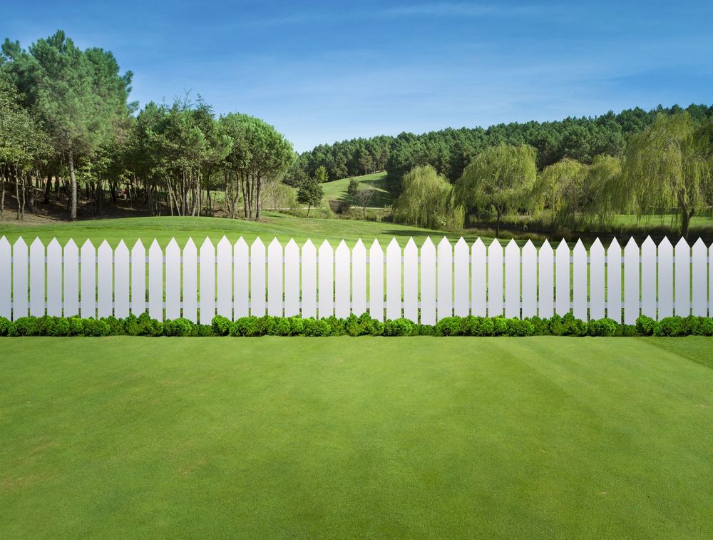 White picket fence bordering a green lawn with trees and a hill in the background under a blue sky.