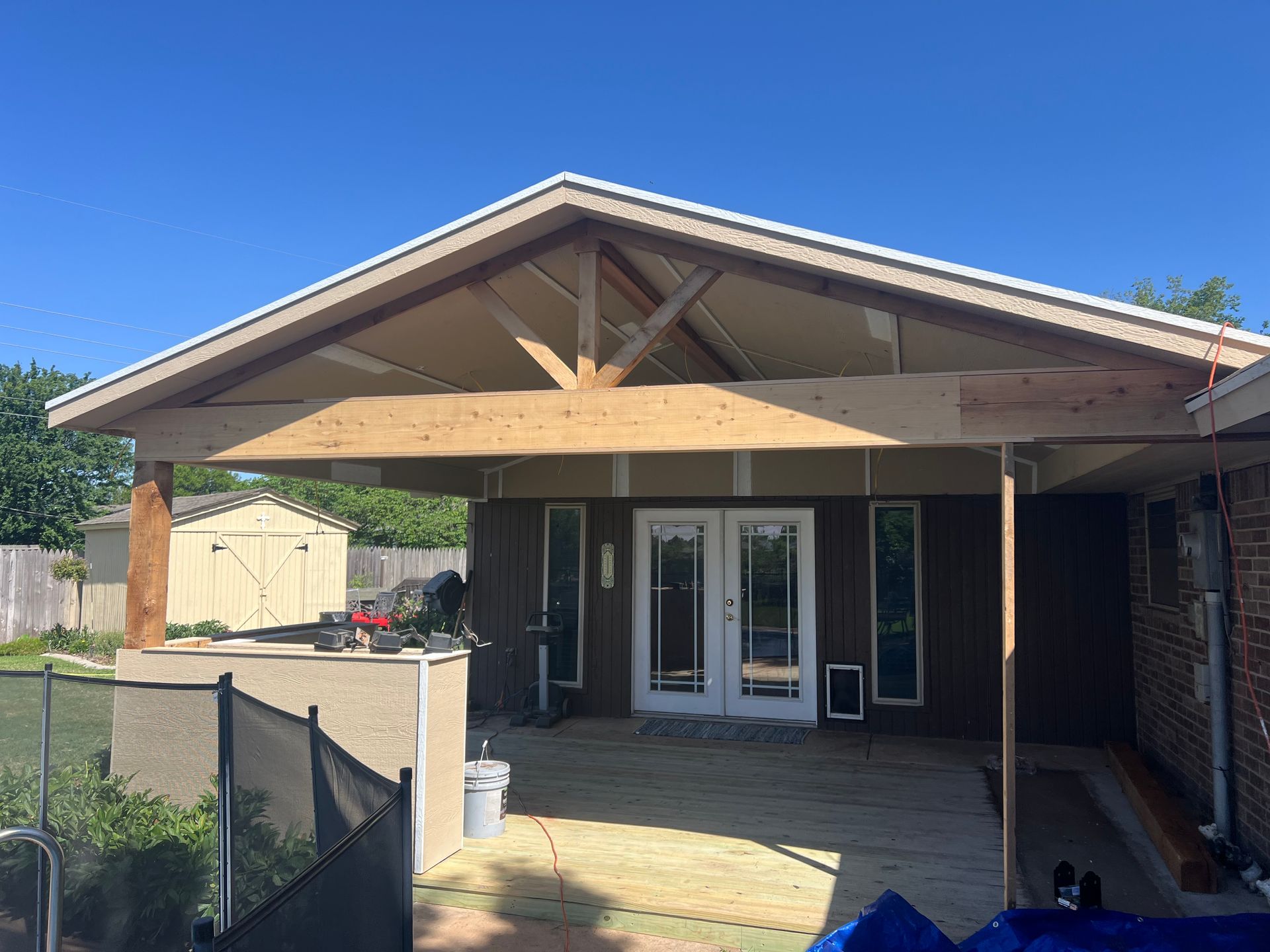 A partially built patio cover with a wooden frame attached to a brown house on a sunny day.
