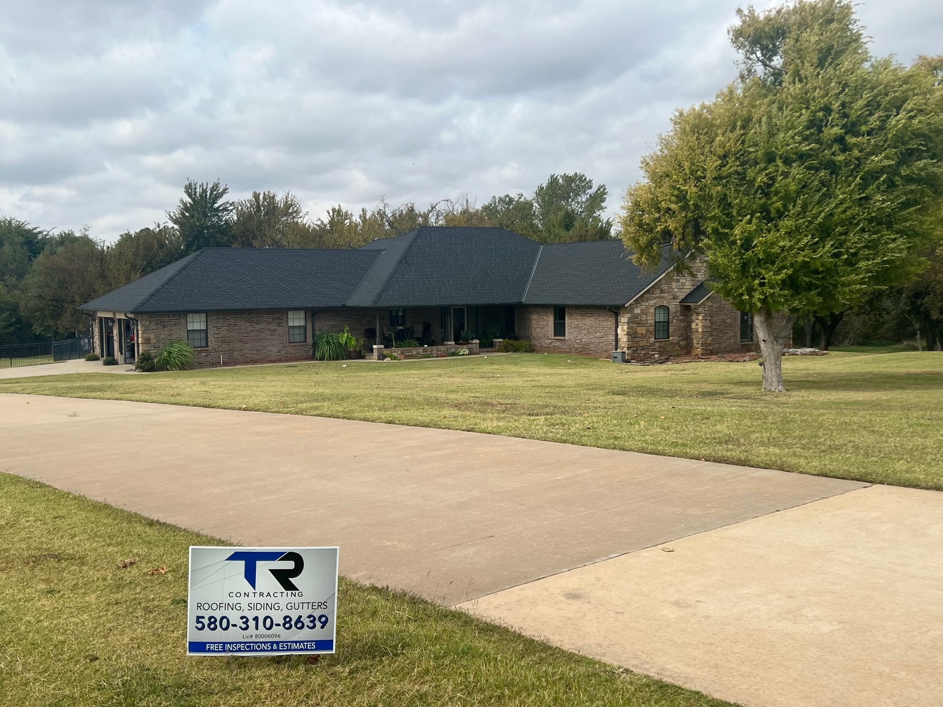 A brick house with a new black roof and a sign in the yard on a cloudy day.