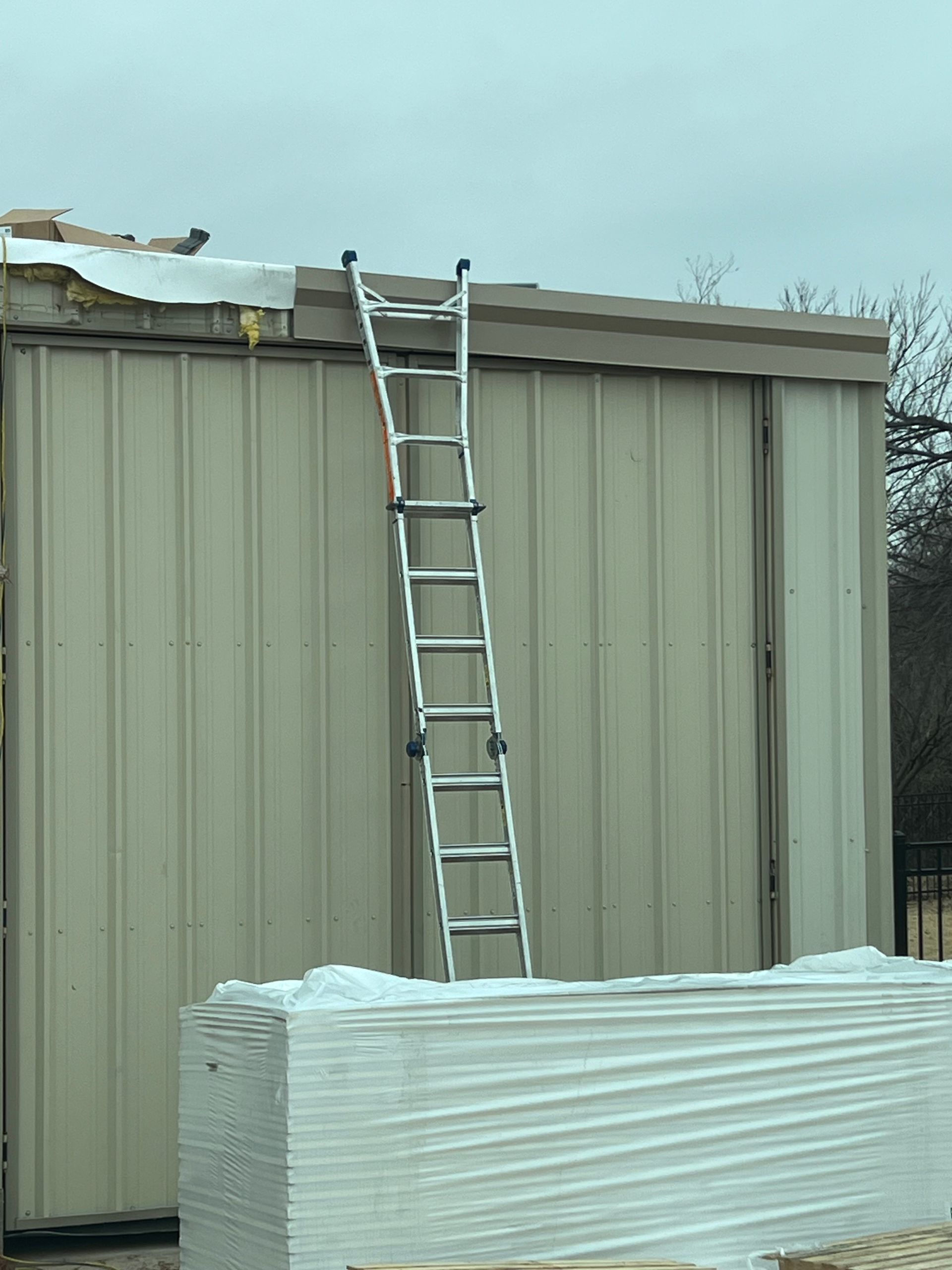 Ladder leans against a tan metal building. Bundles of white boards sit below. Cloudy sky.