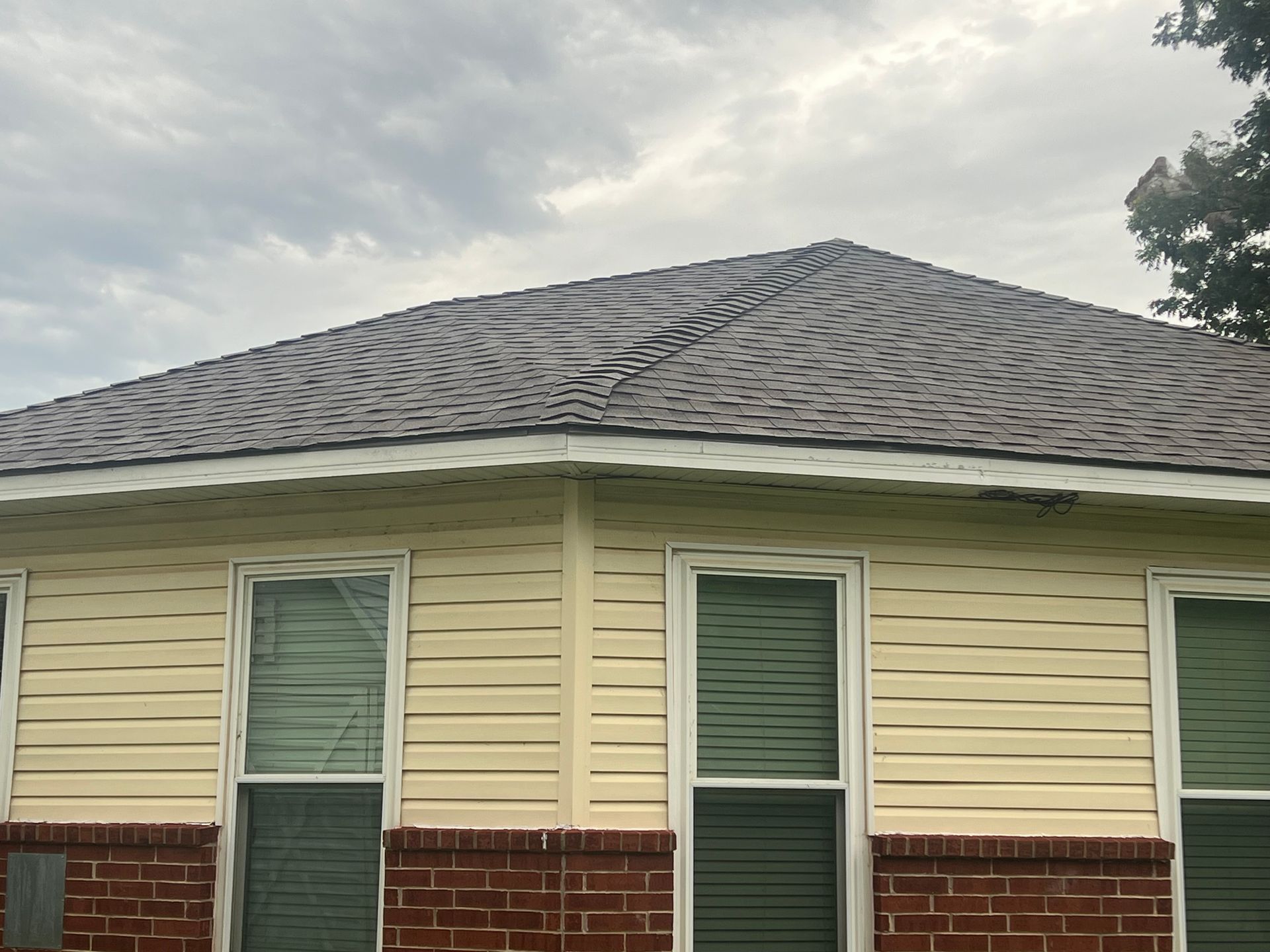 Tan house with brown roof showing weather damage. Windows are visible. Overcast sky.
