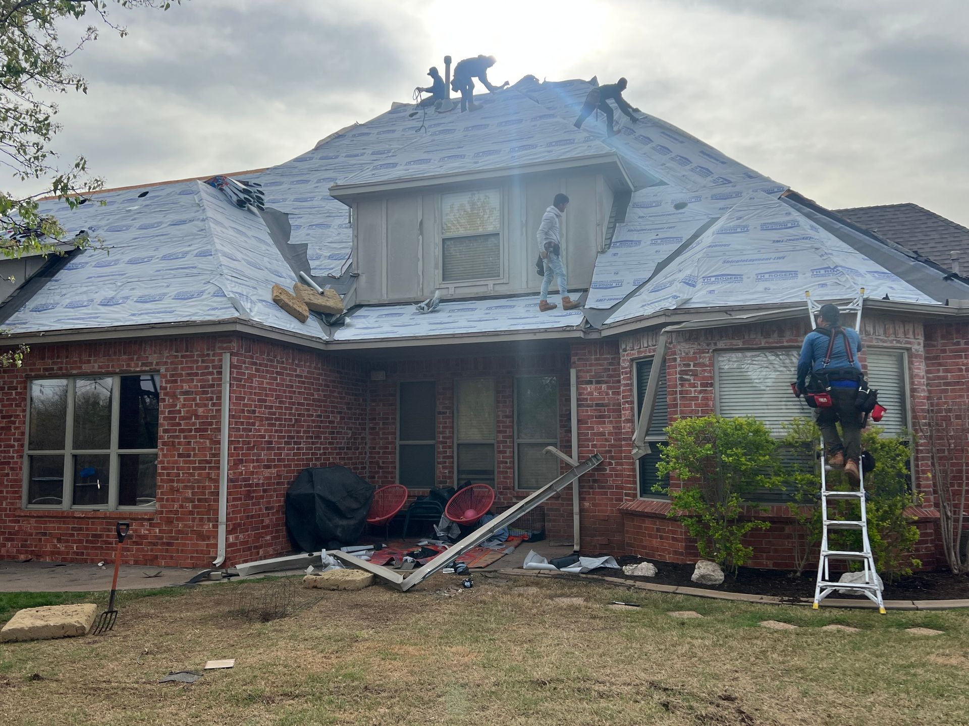 Roofers repairing a brick house, working on the roof with ladders and tools.