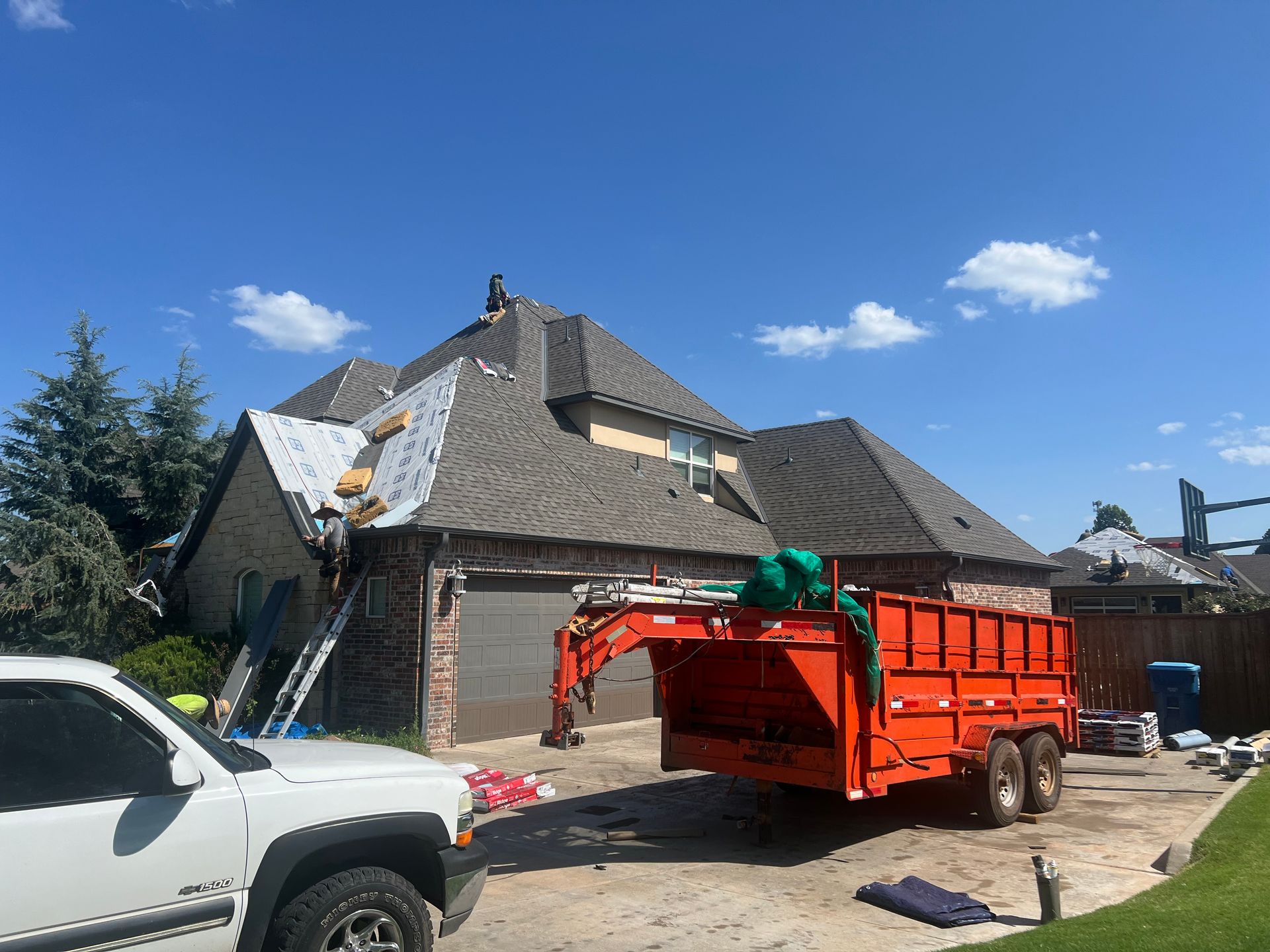 House with roofing repair in progress; workers on roof, orange trailer, truck, blue sky.