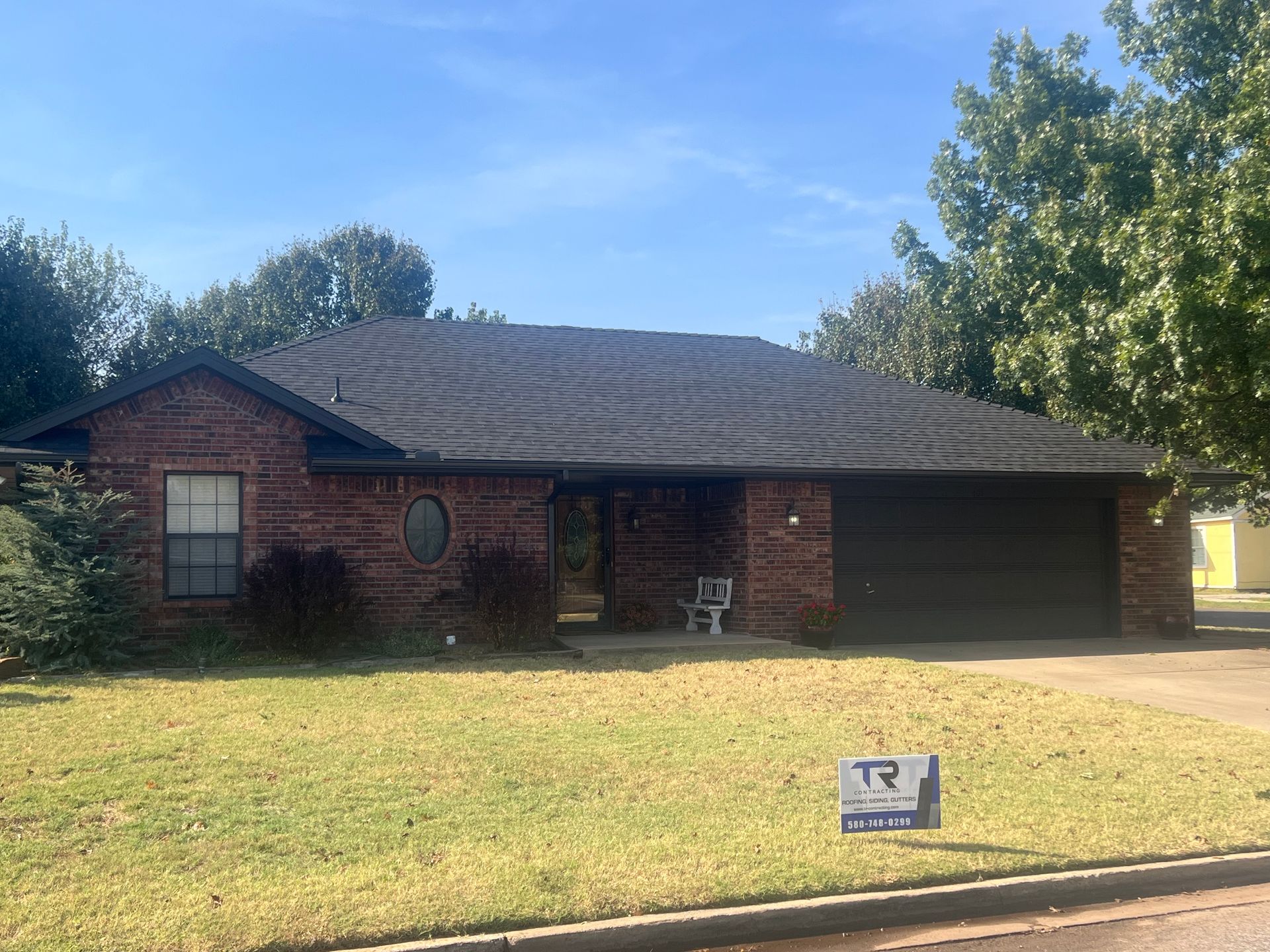 Brick house with black roof and garage, sign in lawn, and trees under blue sky.