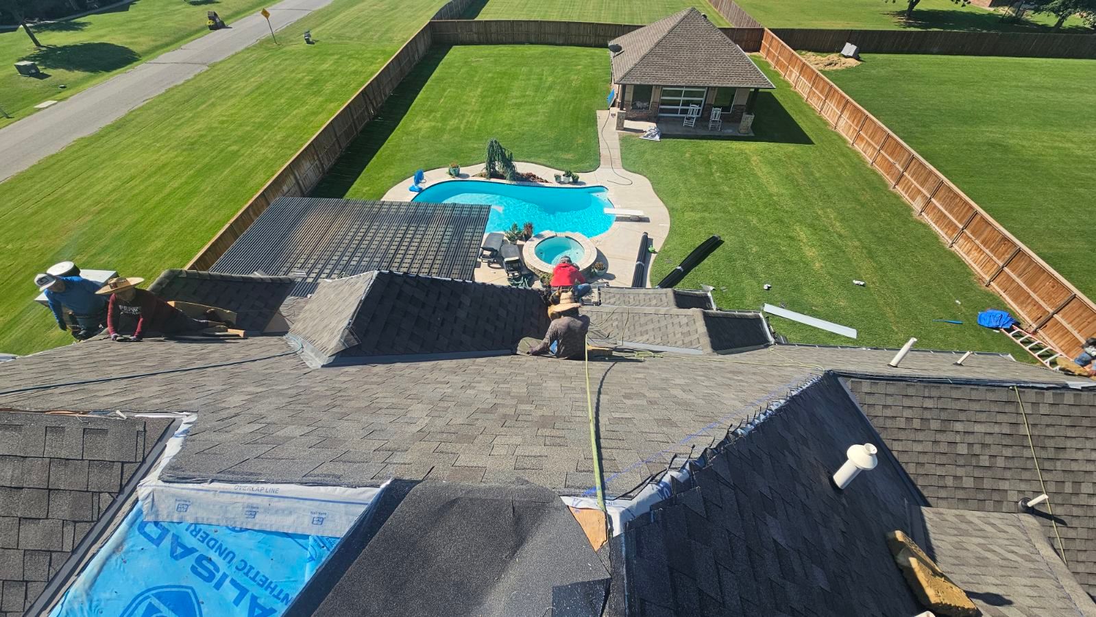 Roofers working on a house roof, pool and gazebo in the background, green lawn, sunny day.