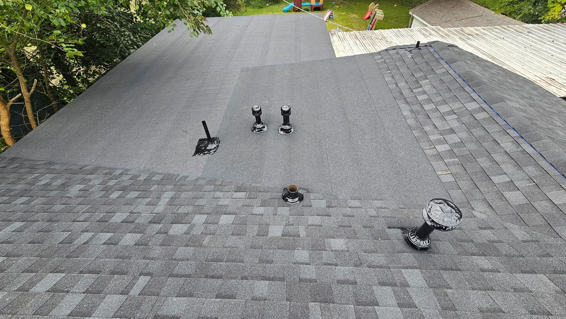 Gray asphalt shingle roof with black vent pipes, seen from above, with trees in the background.