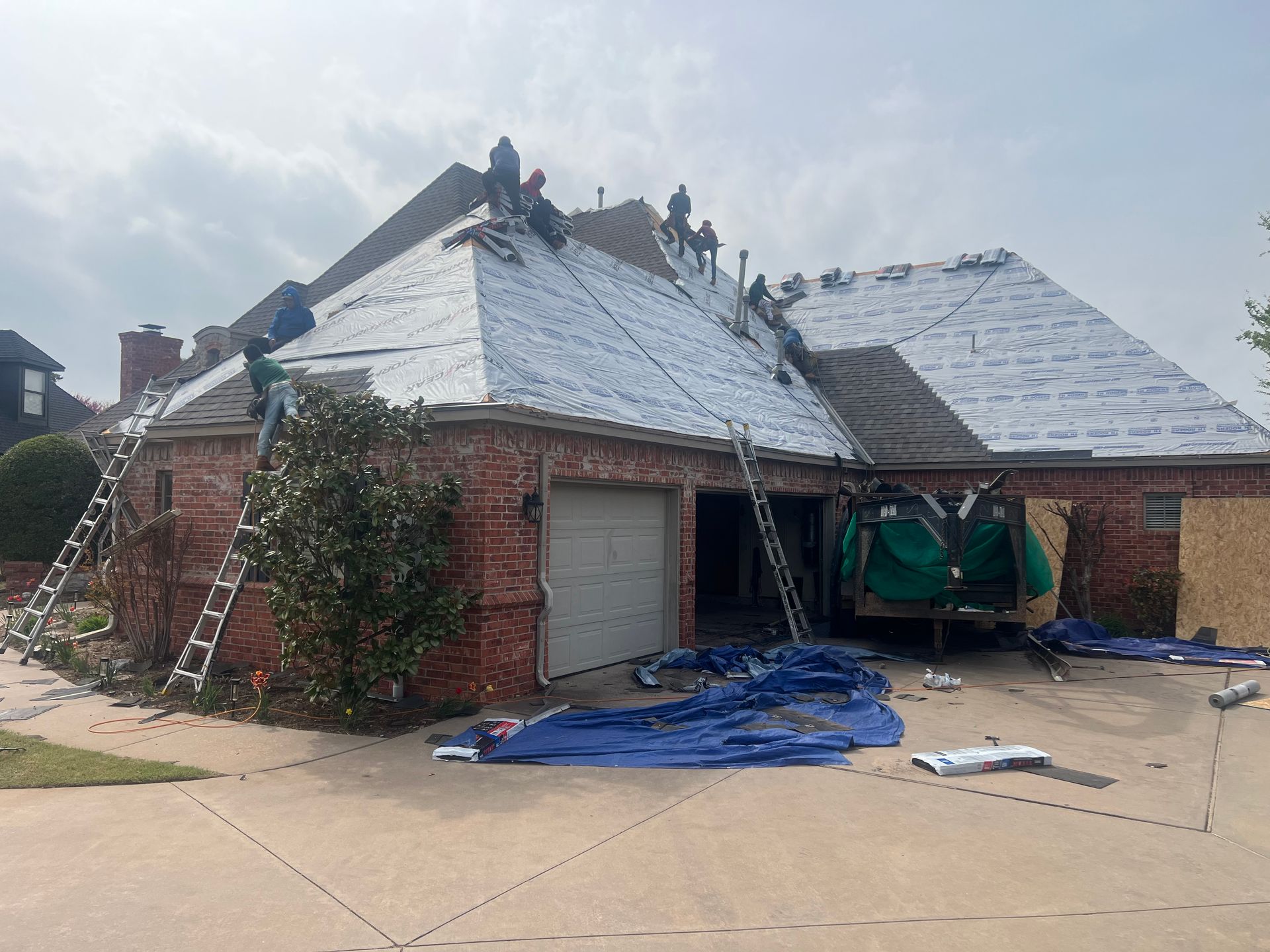 Workers replacing a roof on a brick house. Ladders, tarps, and construction materials are visible.