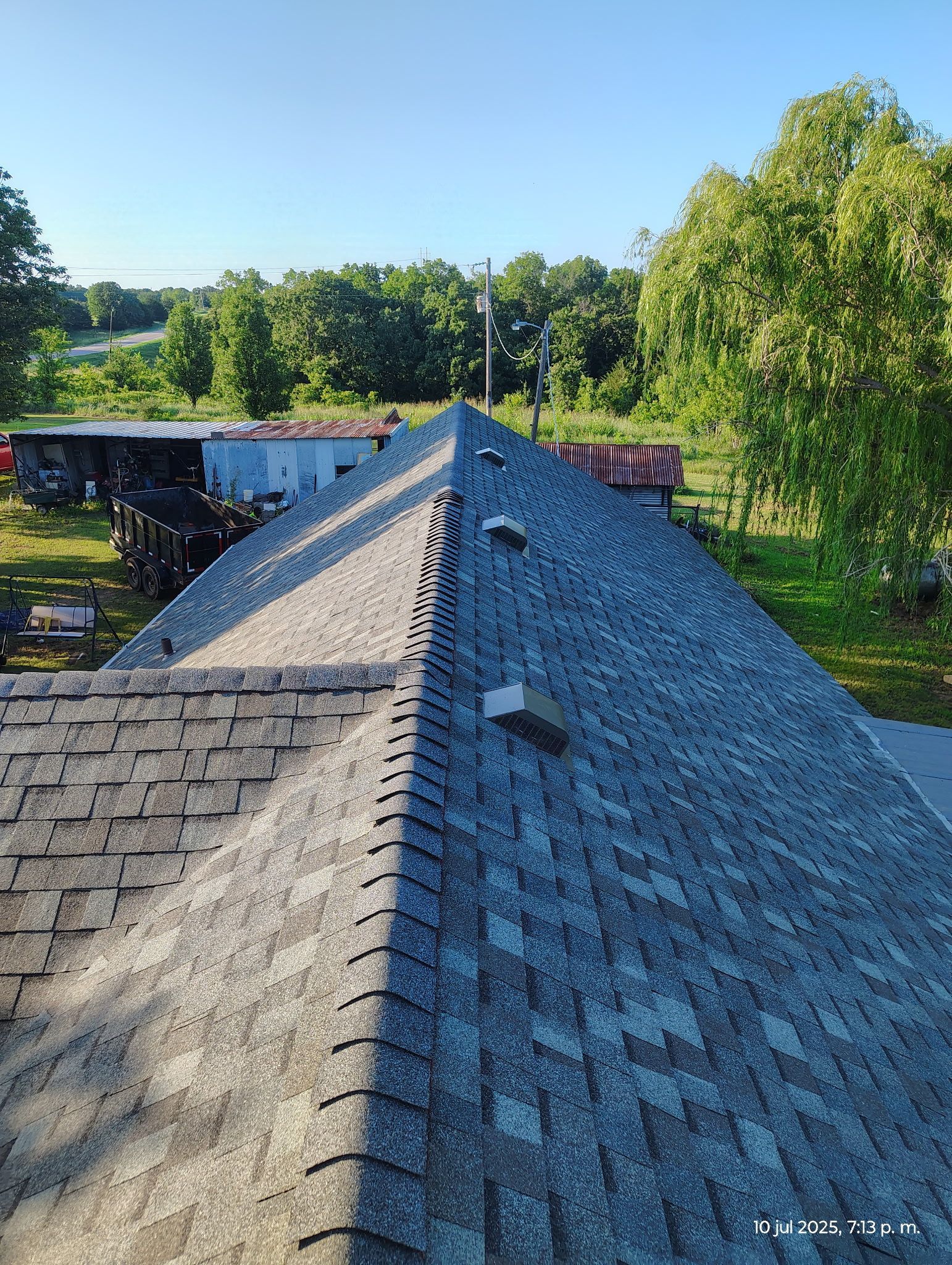 Asphalt shingle roof against a backdrop of trees and a bright blue sky.