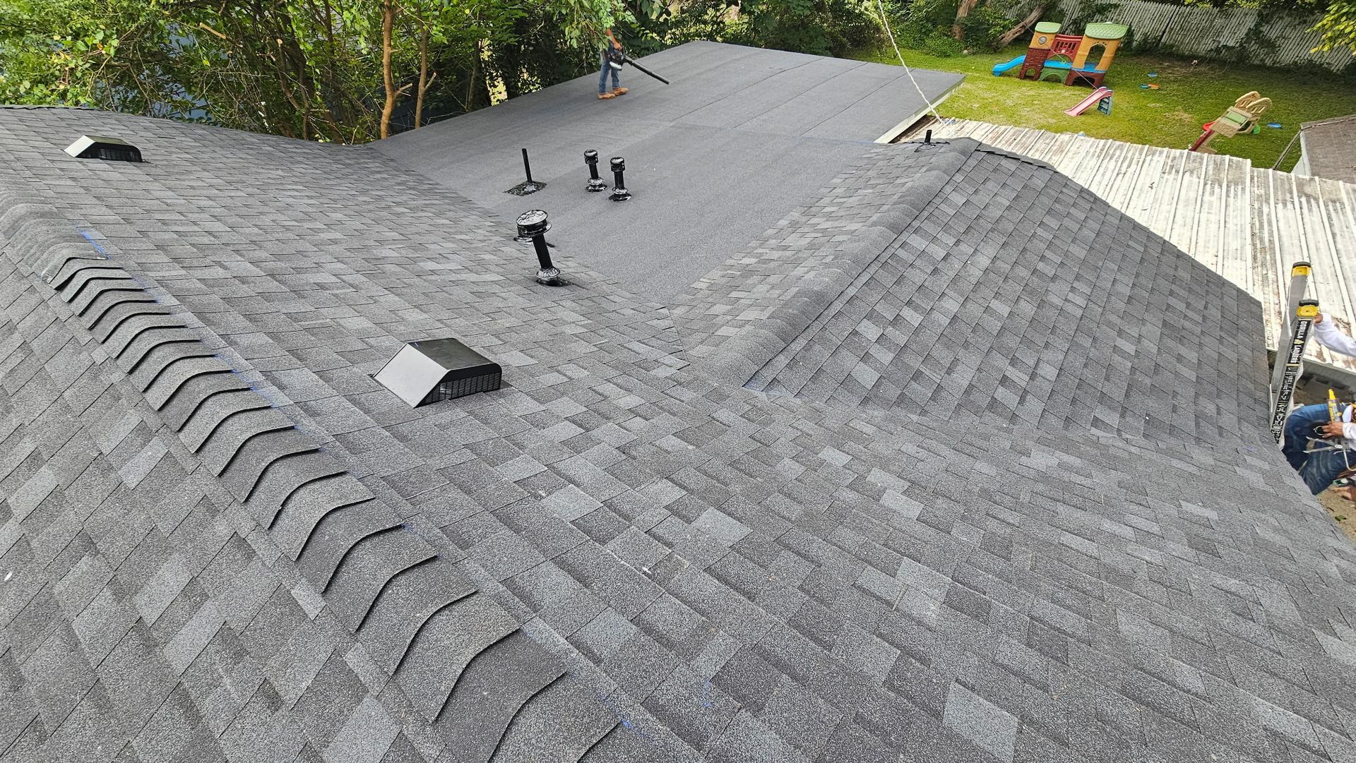Overhead view of a gray asphalt shingle roof with vents and a ridge cap, in a residential setting.
