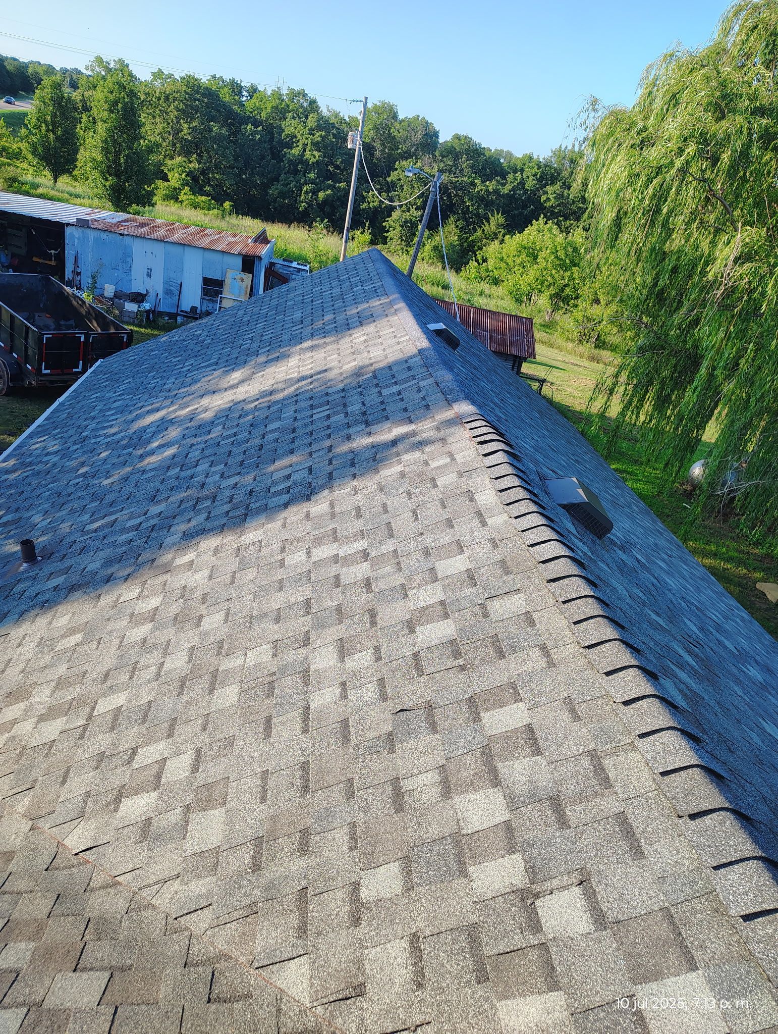 Gray asphalt shingle roof with trees and buildings in the background on a sunny day.