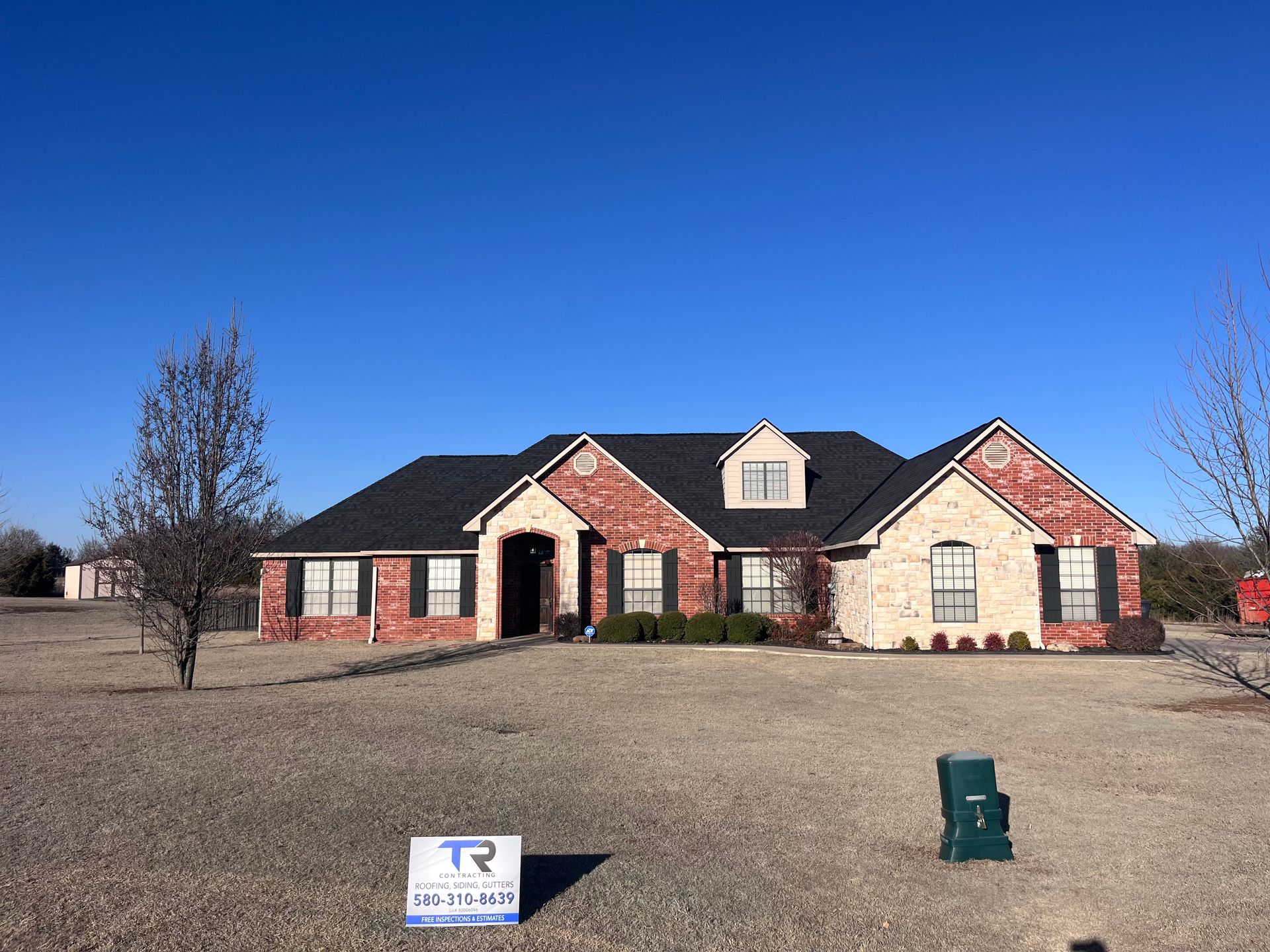 Brick house with black roof under a clear blue sky. A sign and mailbox sit in the yard.