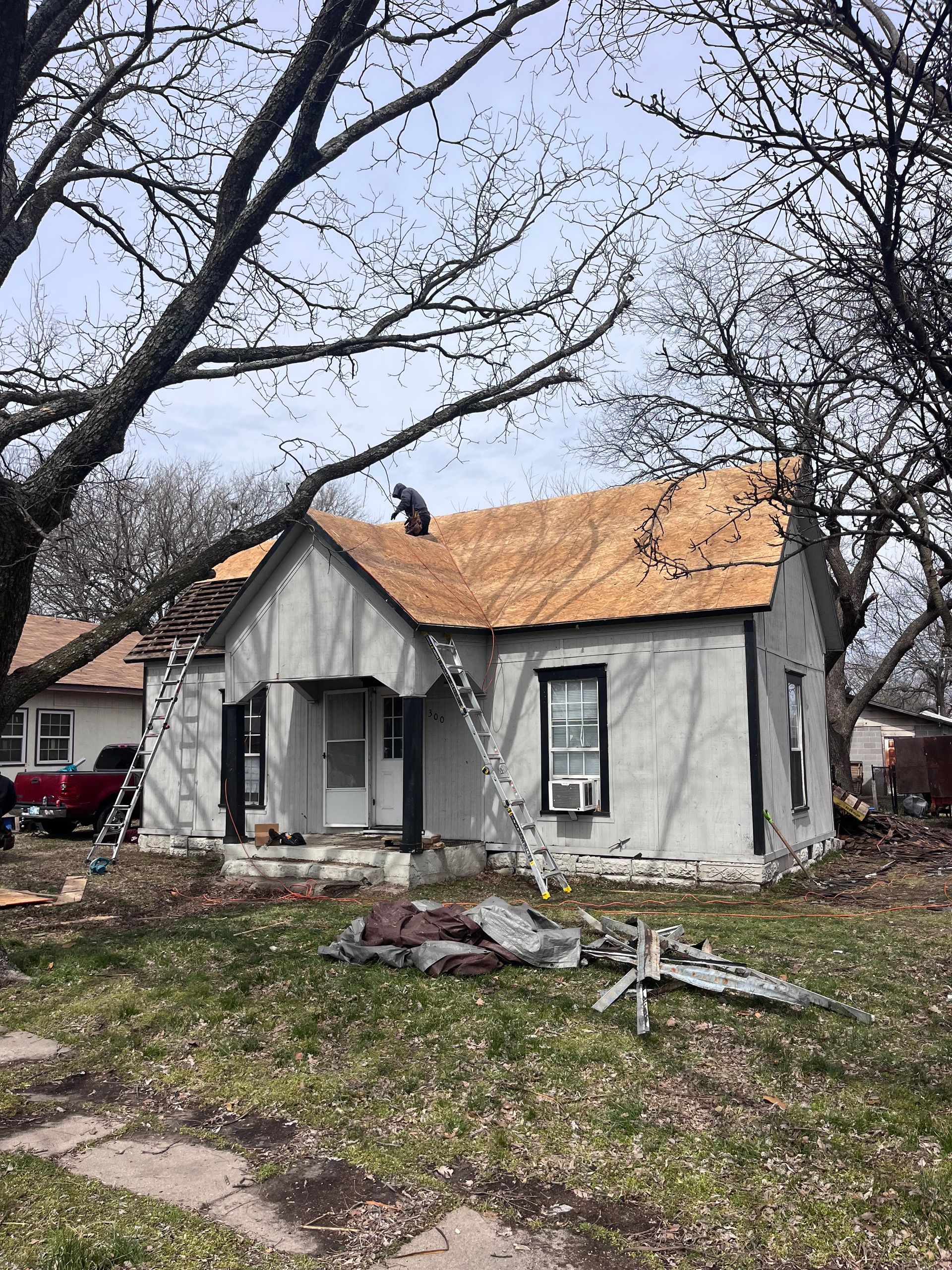 House being re-roofed; person on roof, ladder up, grey house, brown roof, green grass, overcast sky.