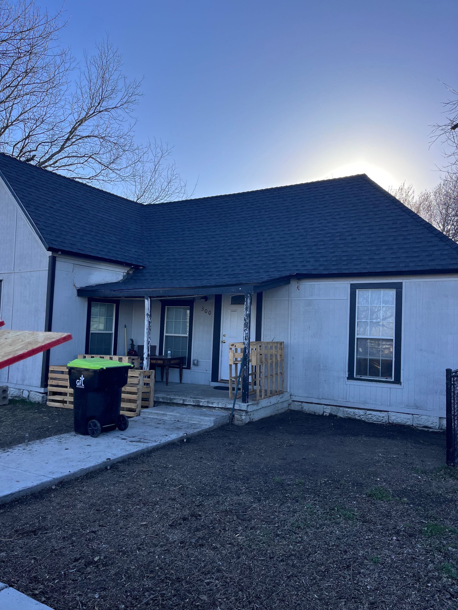 Small, white house with dark roof, porch, and trash cans in front. Clear sky.