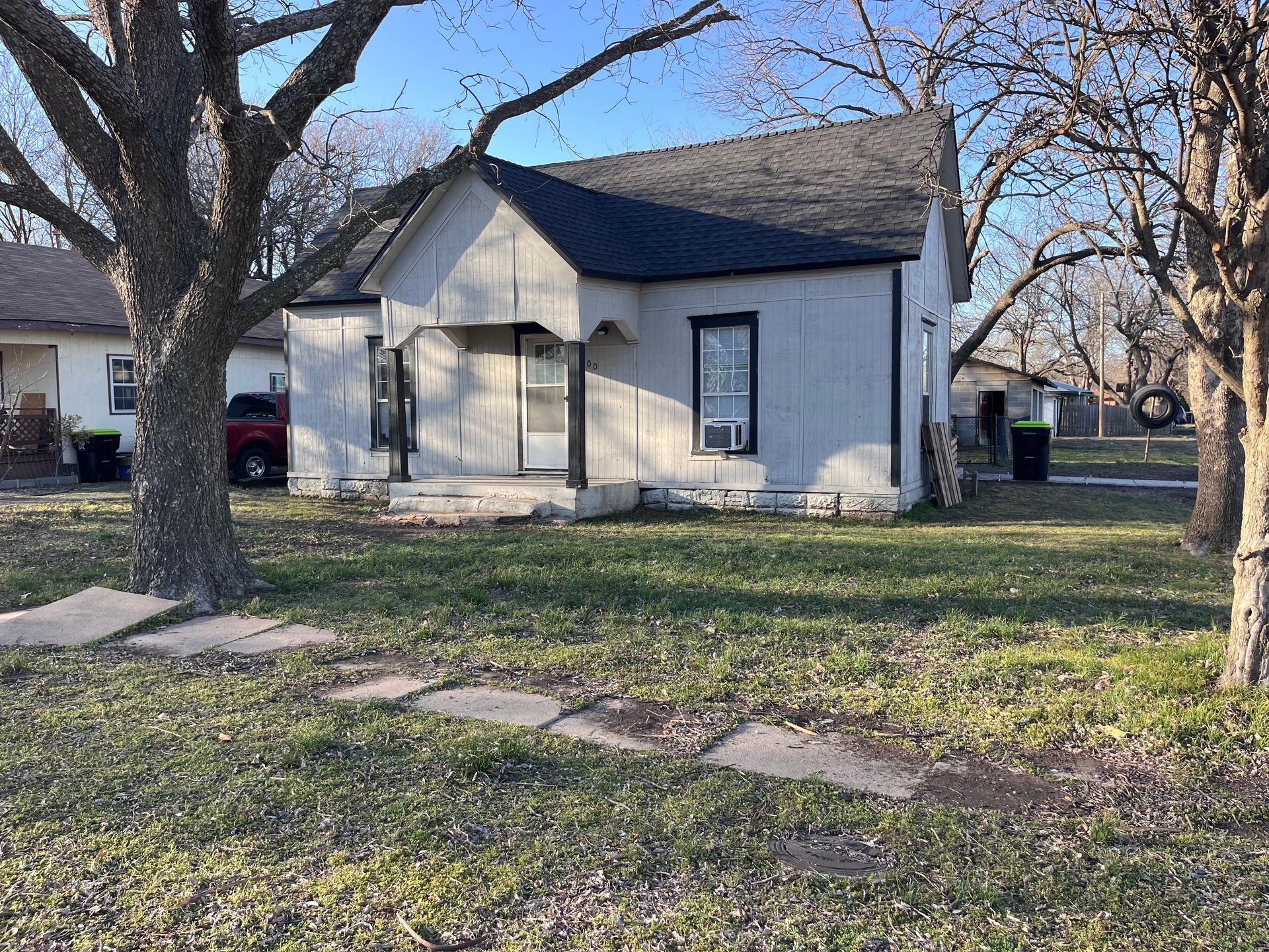 Small, light-colored house with black roof and a tree-lined yard.