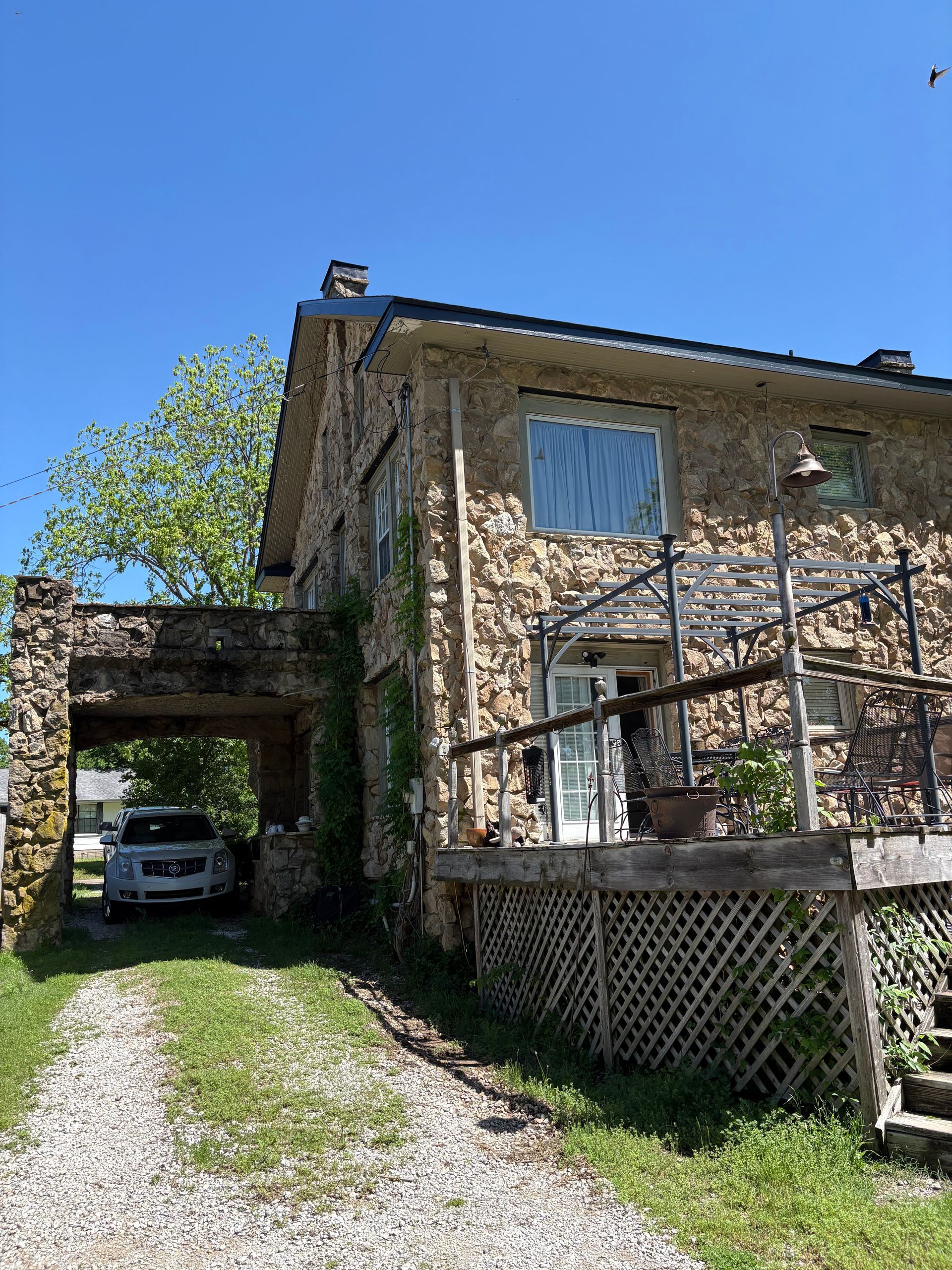 Stone house with gravel driveway, car parked under a stone archway, wooden deck, sunny day.