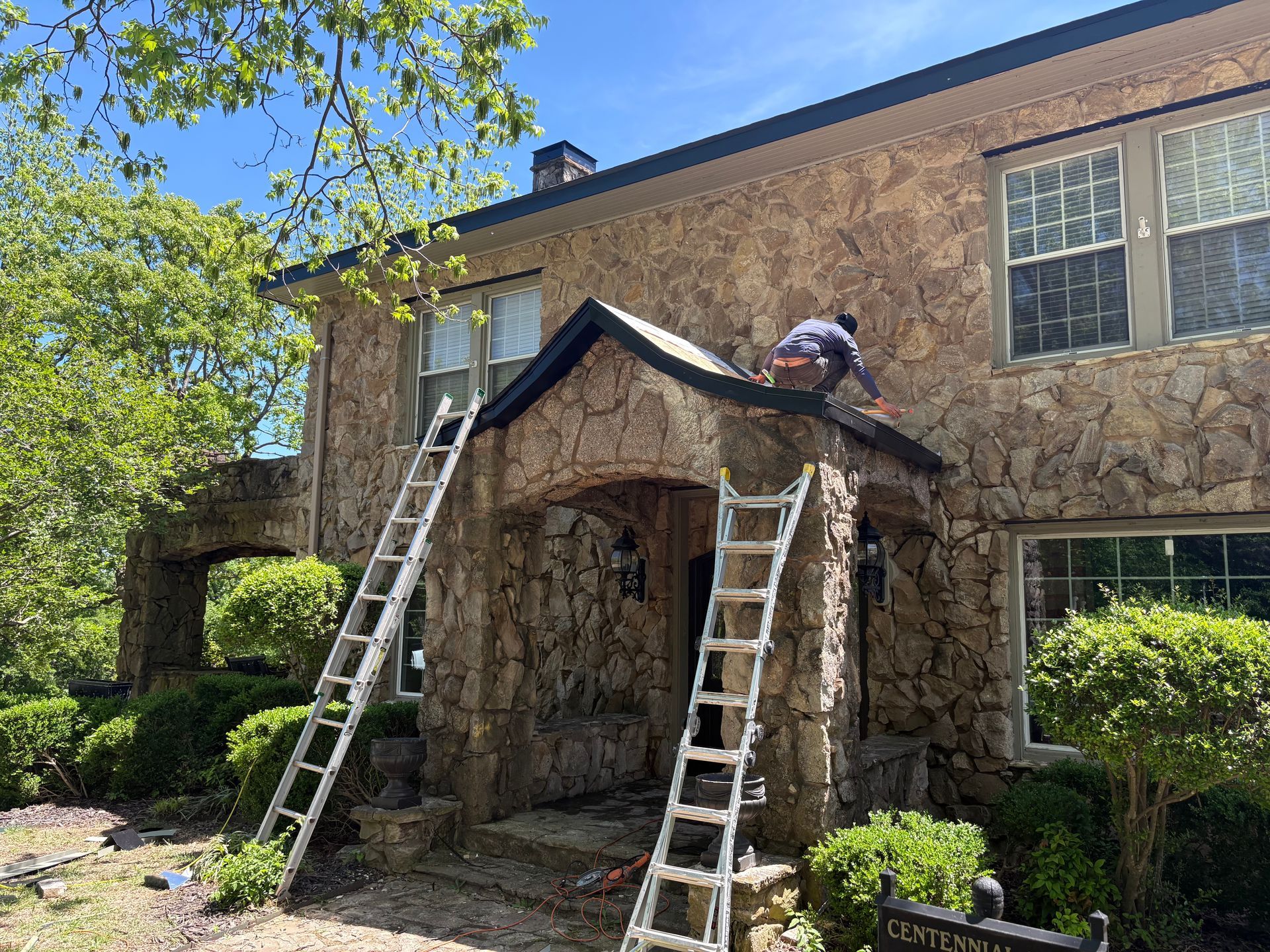 Person on a roof working, two ladders, stone house with arched entrance, trees, and blue sky.
