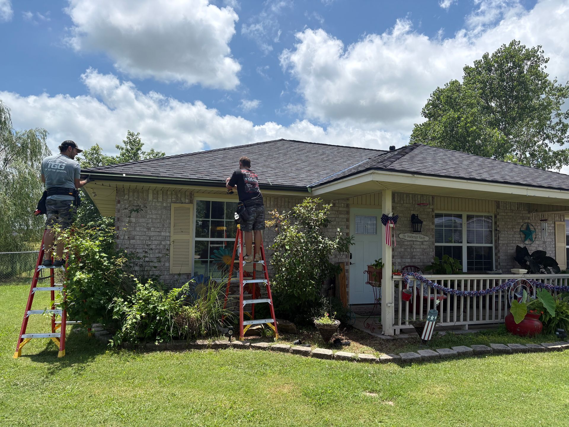 Two people on ladders cleaning a roof, sunny day, house with landscaping.