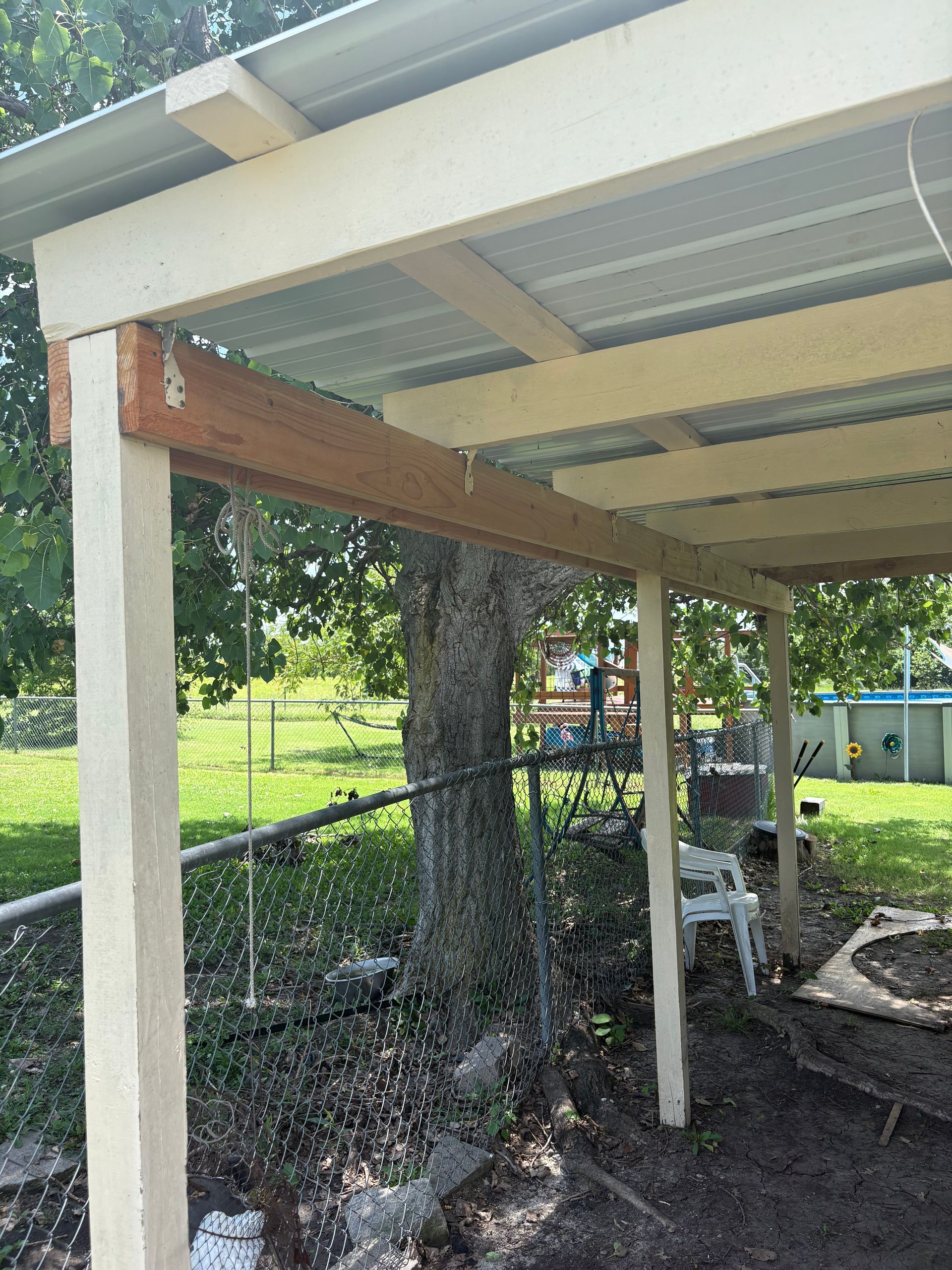 Carport with white posts, metal roof, and a wooden beam, in a yard.