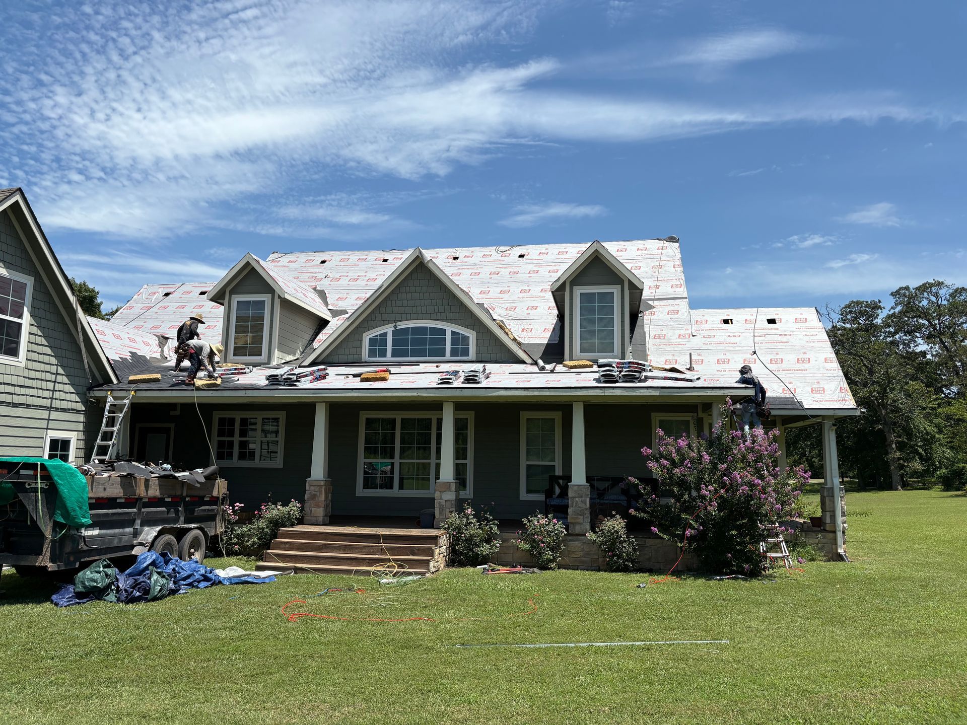 Roofing crew replacing shingles on a house with a green exterior and large windows on a sunny day.