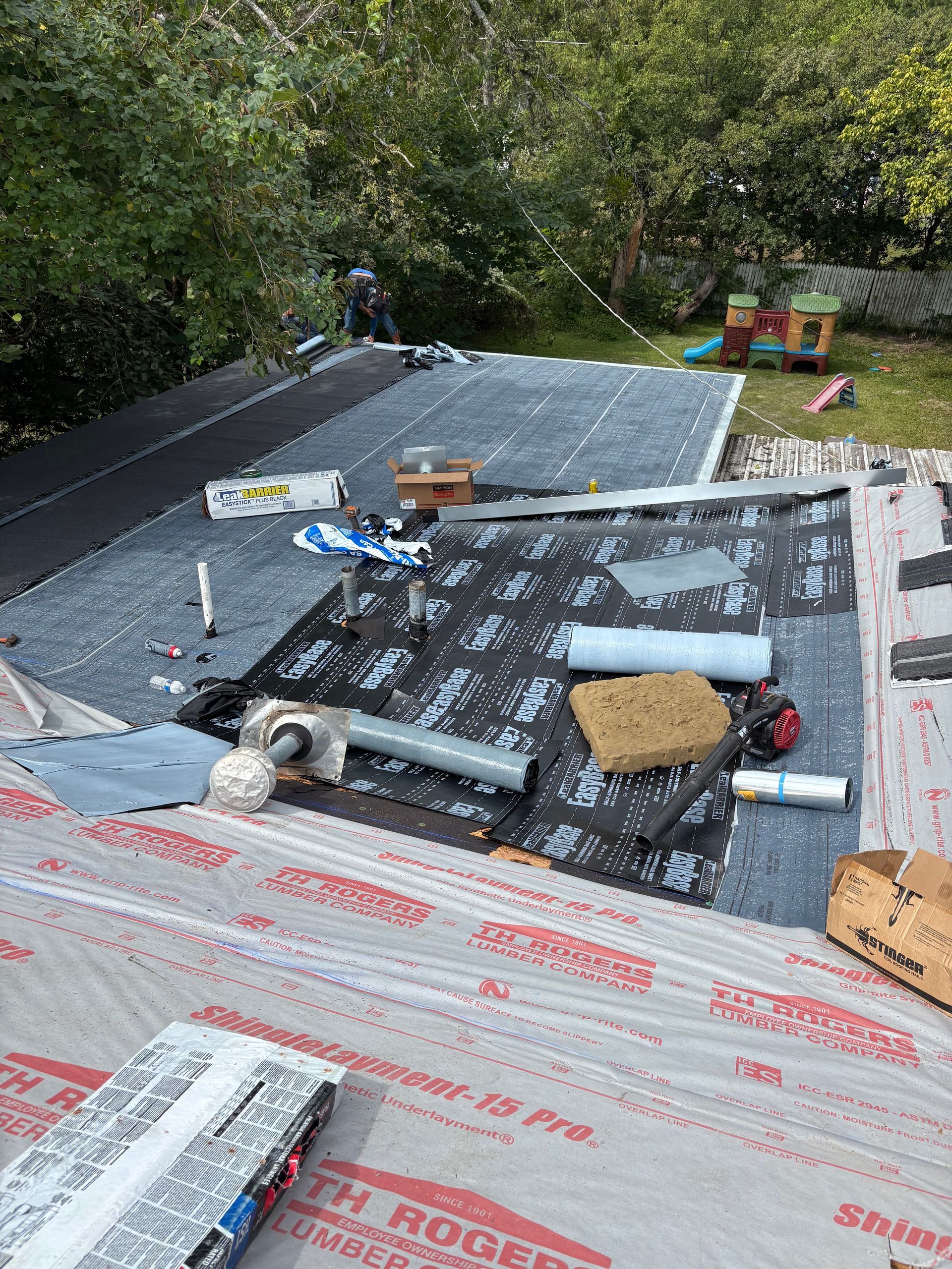 Roofer working on a flat roof, applying black roofing material; tools and supplies are scattered around.