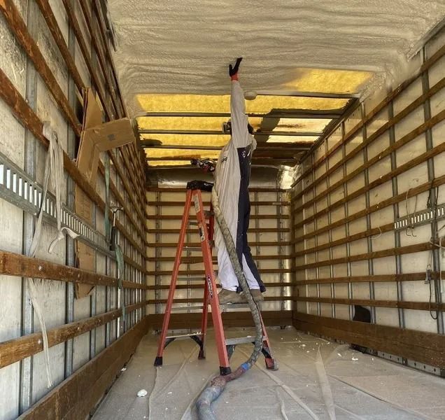Man spraying insulation inside a truck, standing on a ladder. Walls and ceiling covered in foam.