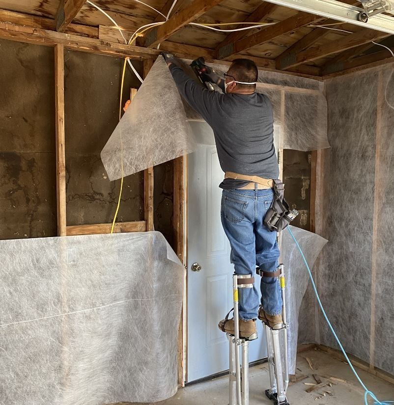 Man on stilts installs insulation in a room with exposed framing and wiring.