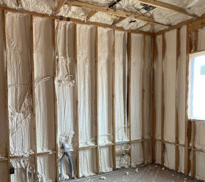 Interior wall of a room, covered with light-colored spray foam insulation, revealing wood framing.