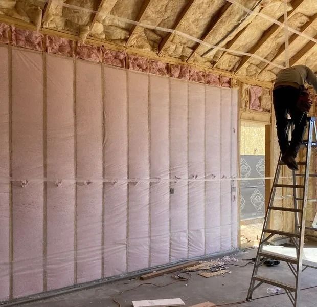 Worker installs pink insulation on a wall inside a building, with a ladder and exposed ceiling beams.