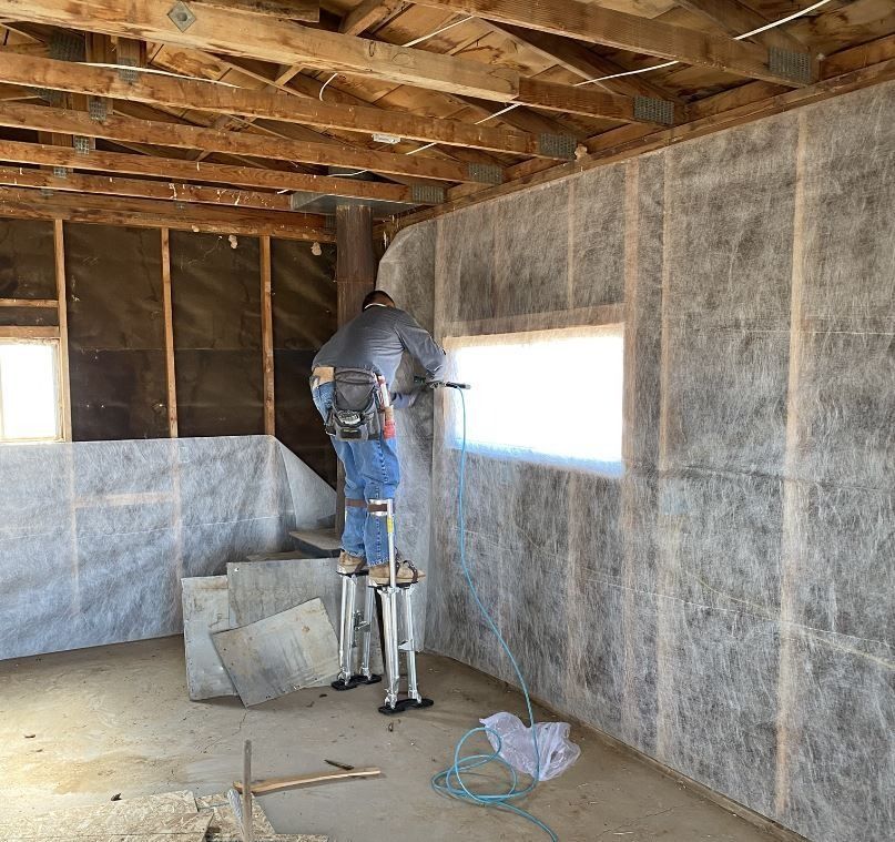 Man on stilts works on wall inside a building, near a window.