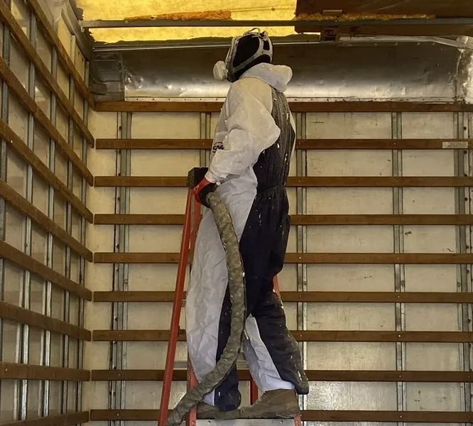 Person in protective suit on a ladder, spraying insulation inside a metal-framed wall.