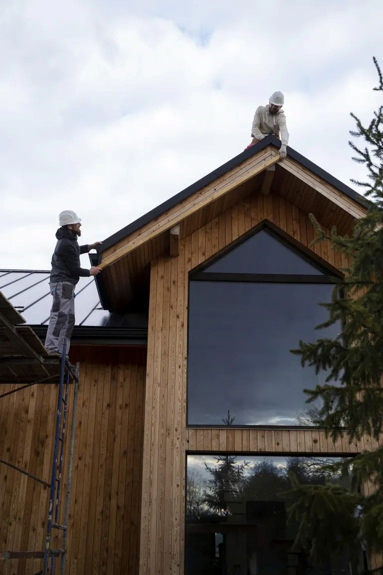 Two workers on a wooden roof, installing something. Large window visible on a wood-clad building.