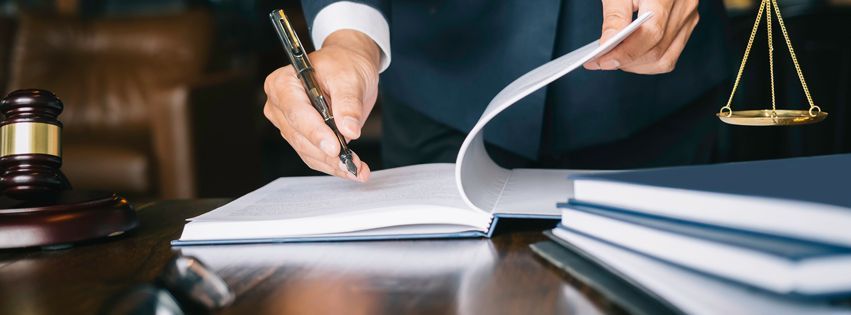 Person in a suit writing on a document at a desk with a gavel, books, and scales of justice.