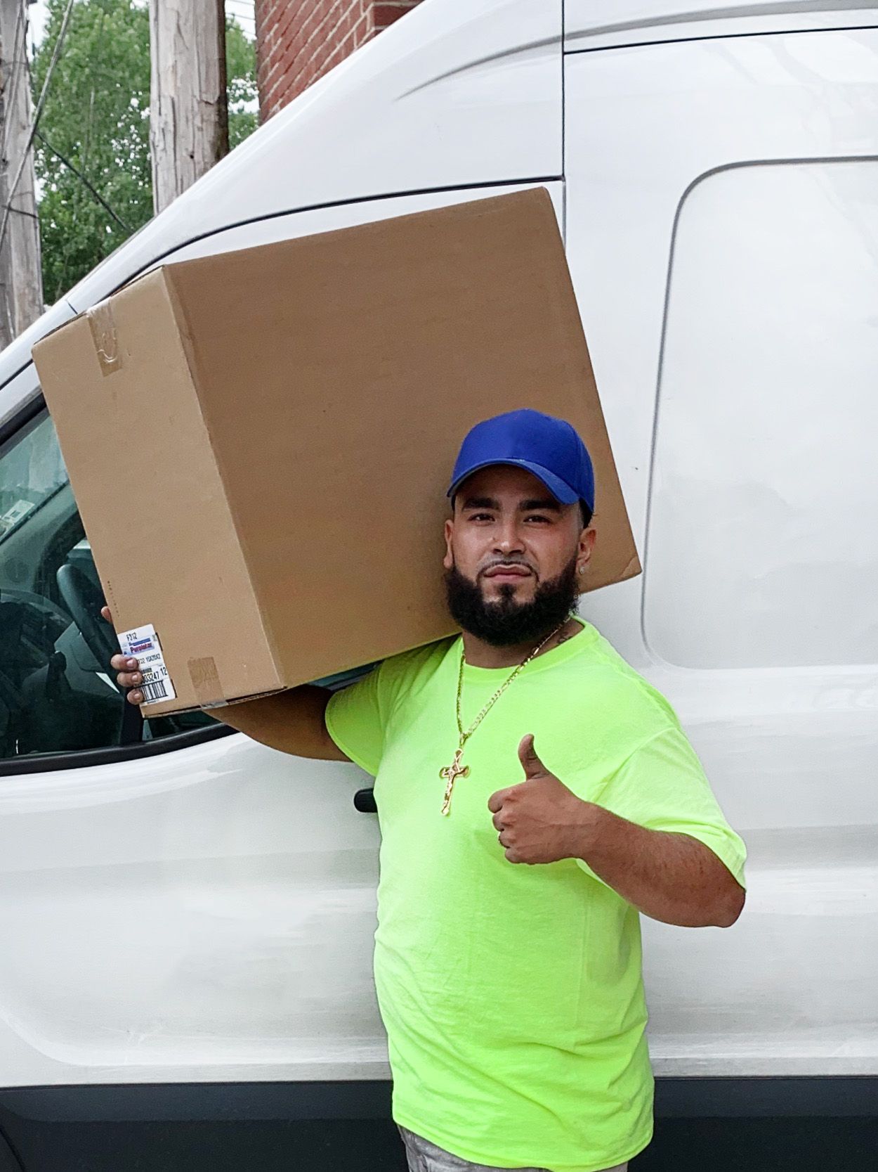 A man in a neon green shirt is carrying a large cardboard box on his shoulder.