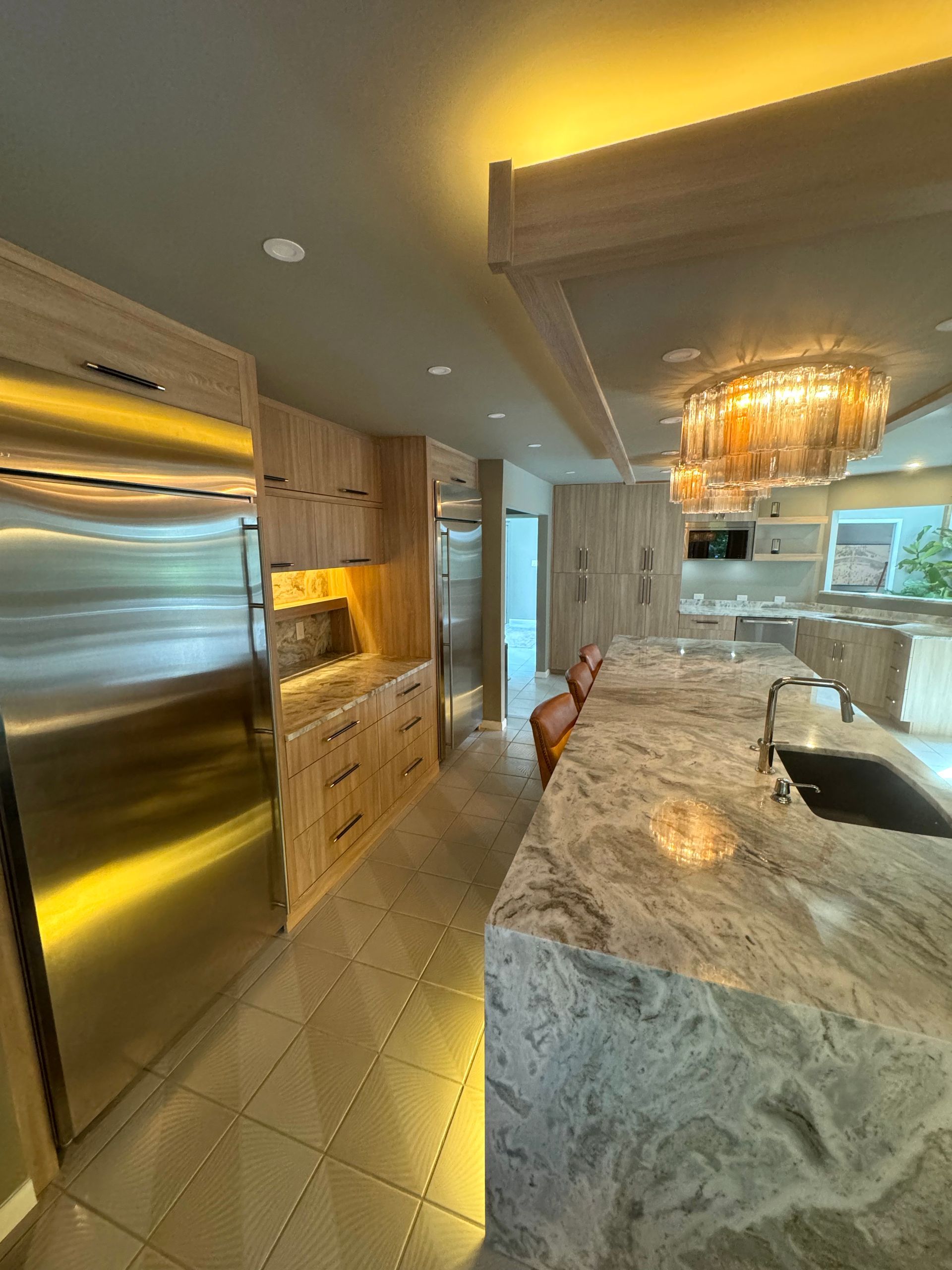 Modern kitchen with stainless fridge, marble island, wood cabinetry, and a chandelier over the counter.