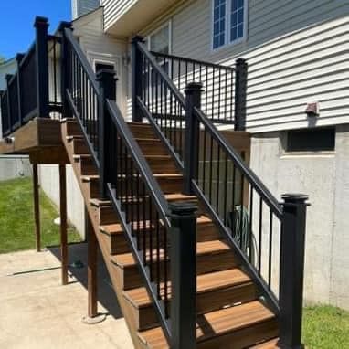 A set of wooden stairs leading up to a deck with a black railing.