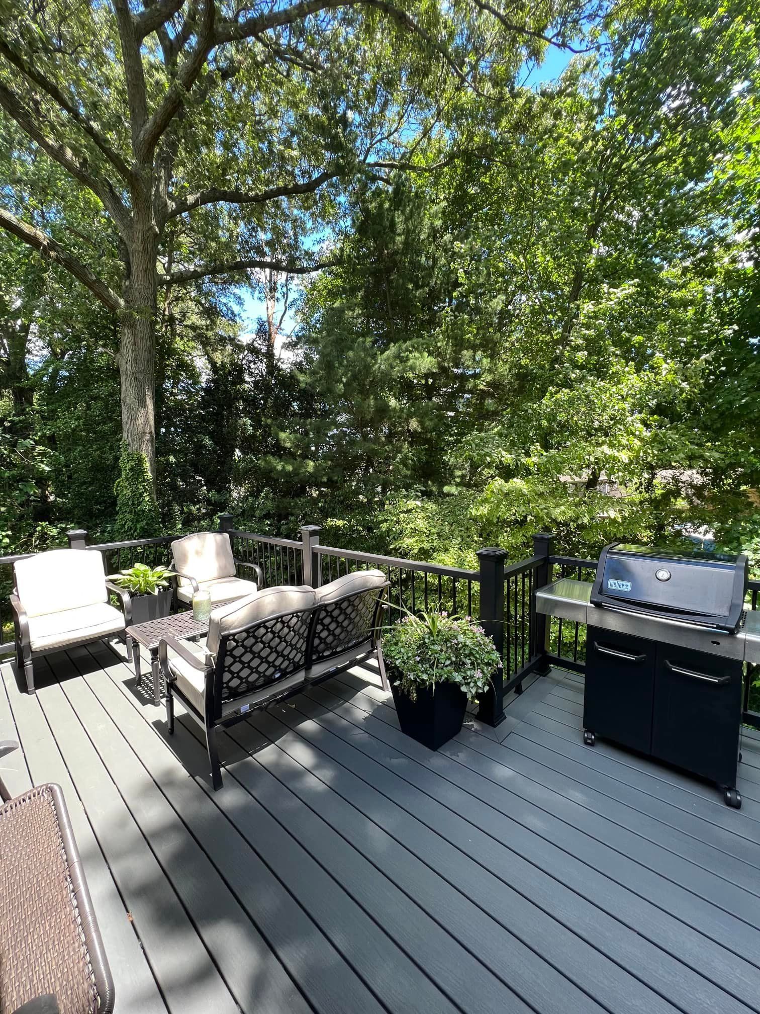 A deck with a couch , chairs , and a grill surrounded by trees.