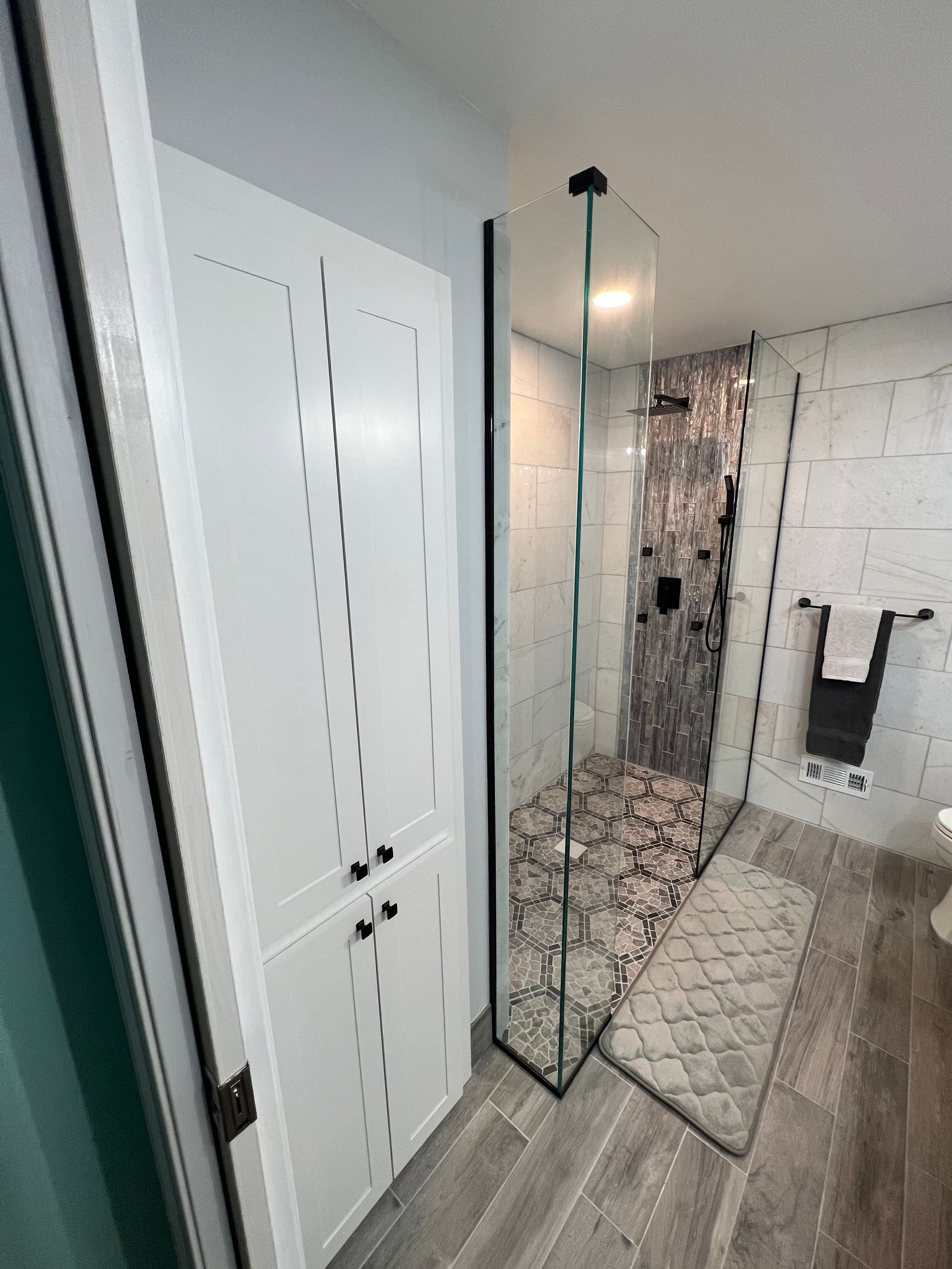 A bathroom with a glass shower, white cabinets, stone floor, and gray tiled walls.