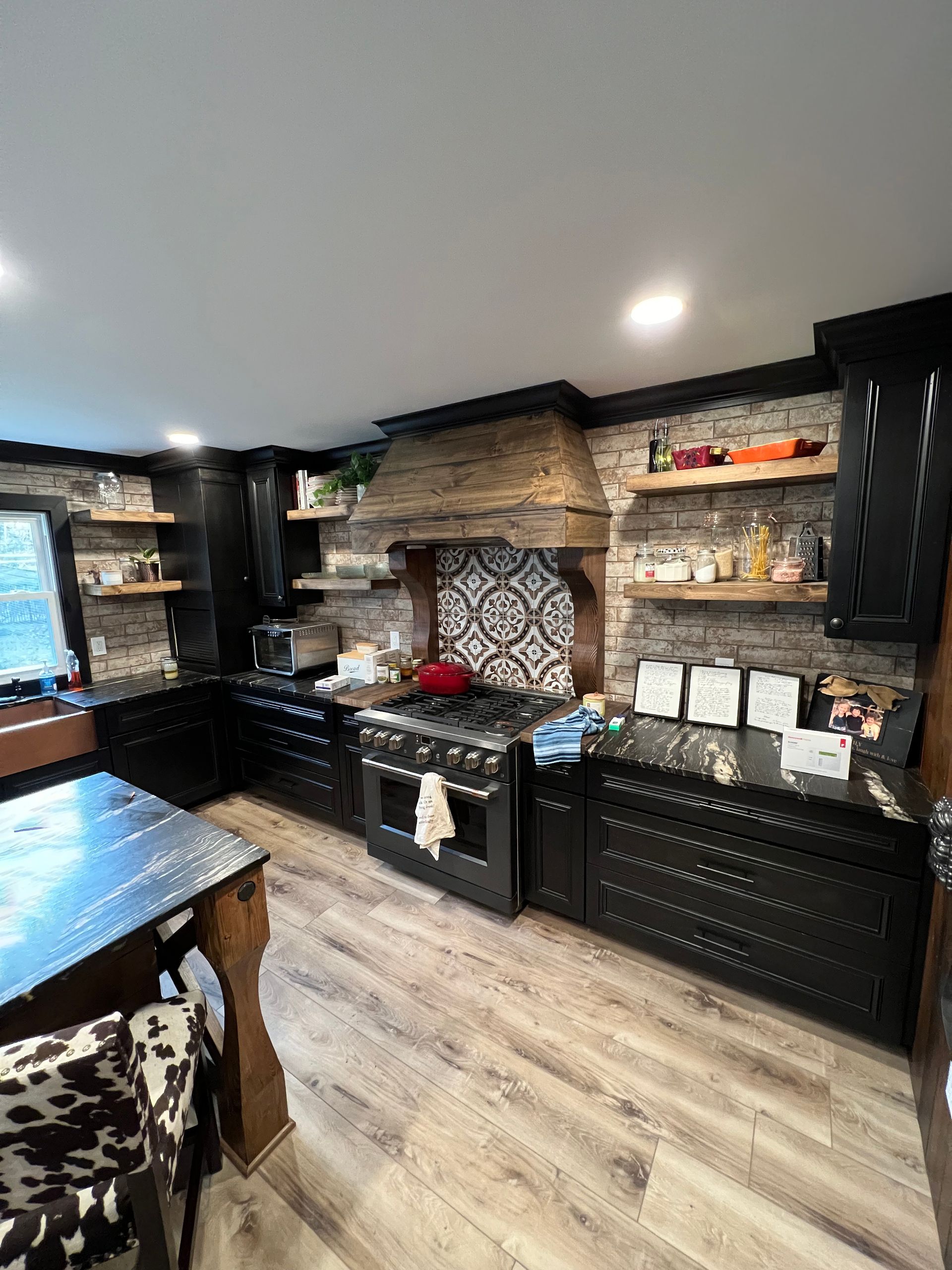 Black kitchen with brick wall, black cabinets, wood shelves, and a wooden hood above the stove.