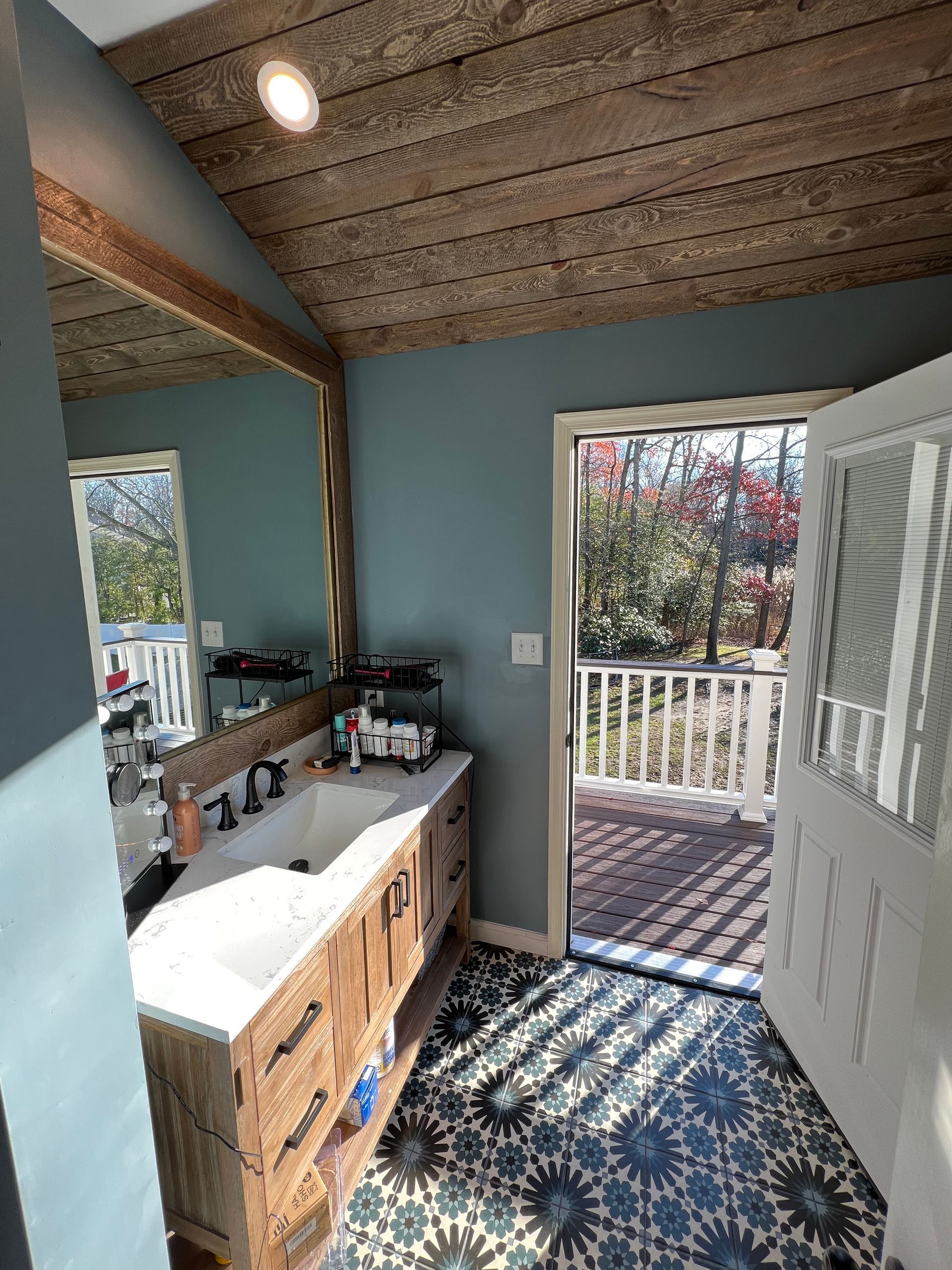 Bathroom with blue walls, wood ceiling, and patterned tile floor opens to a porch with blooming trees.