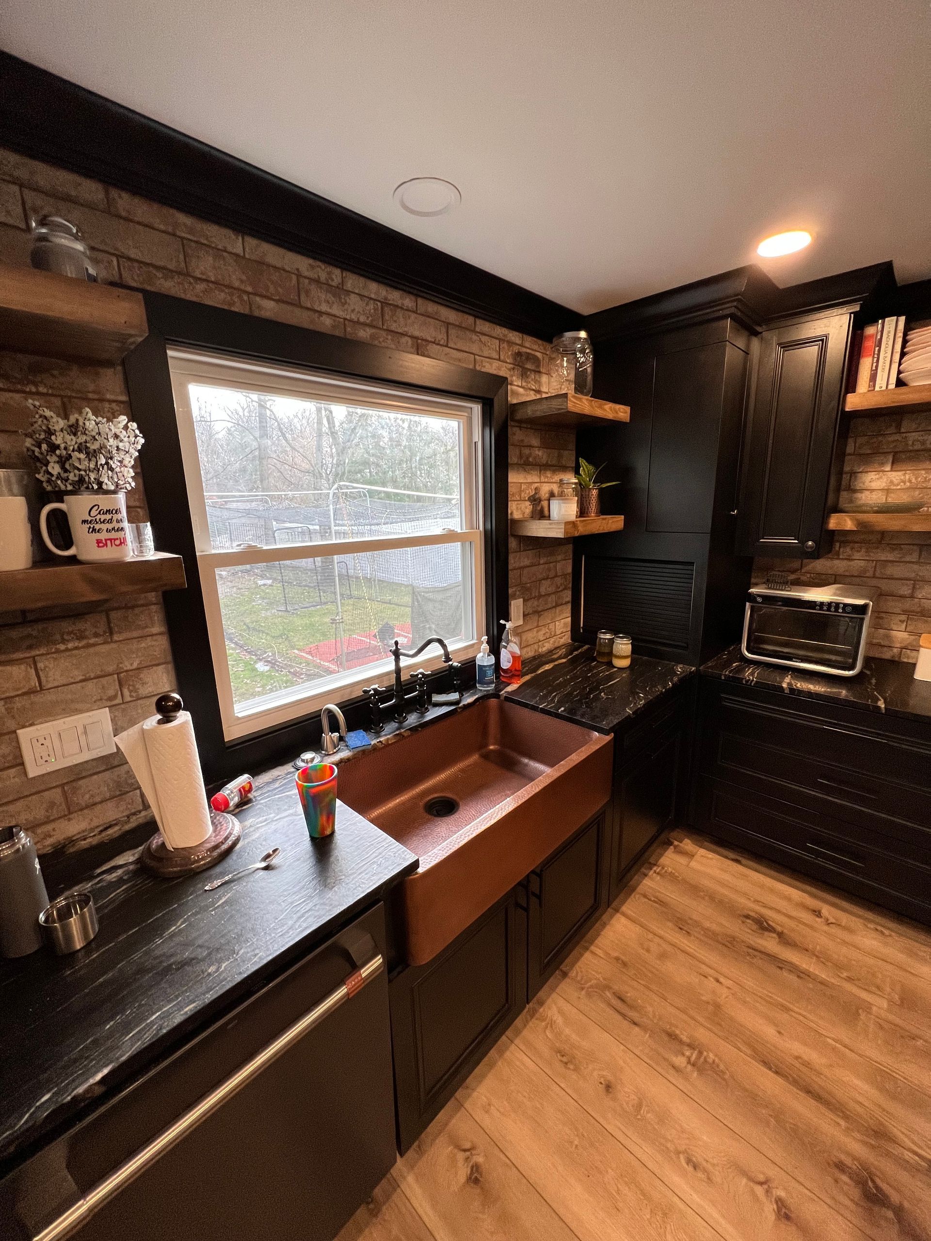 Kitchen with copper sink, black cabinets, brick wall, and wooden shelves.