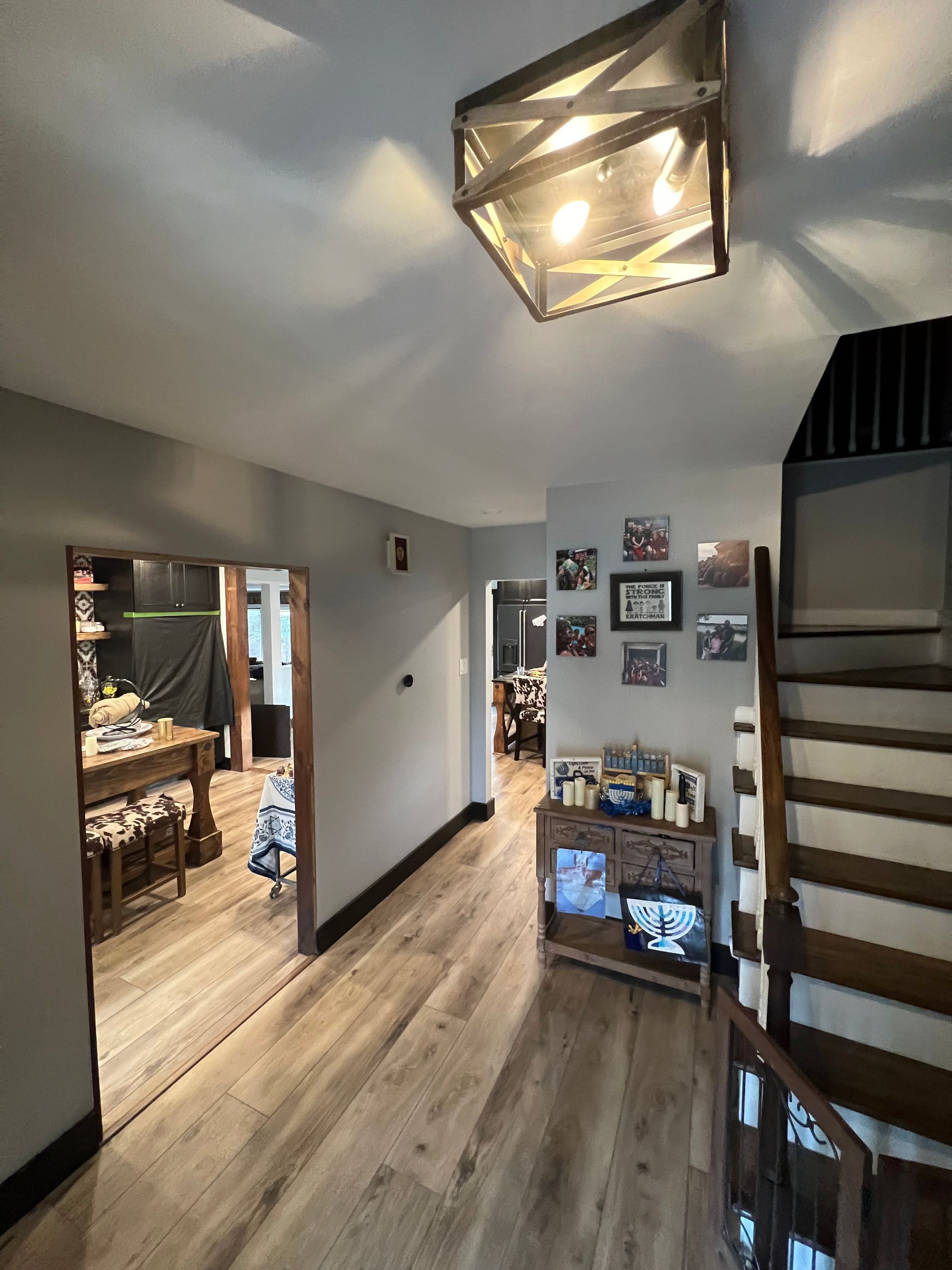 Hallway with wood floors and stairs. Gray walls, rustic light fixture. Kitchen in the distance.