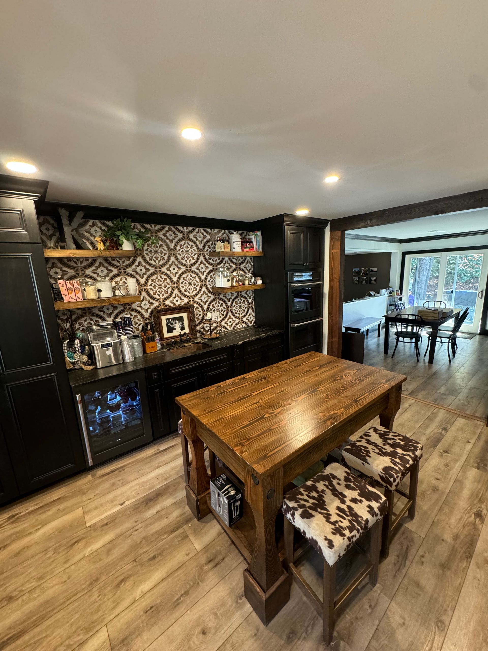 A kitchen with dark cabinets, a wooden island, and cowhide stools, a patterned backsplash, and a dining area visible.