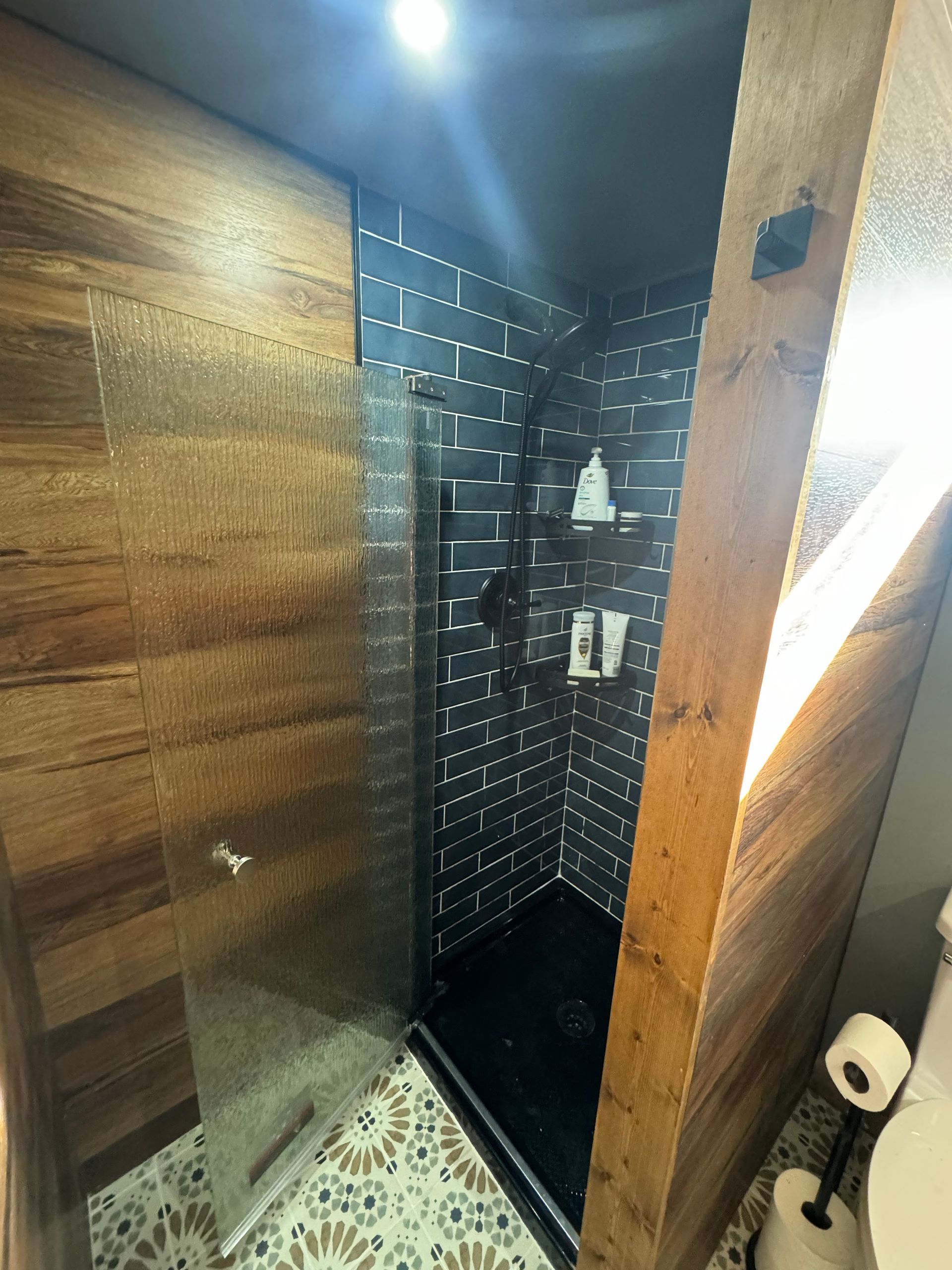 Bathroom with wooden walls, black tiled shower, glass door, and patterned floor.